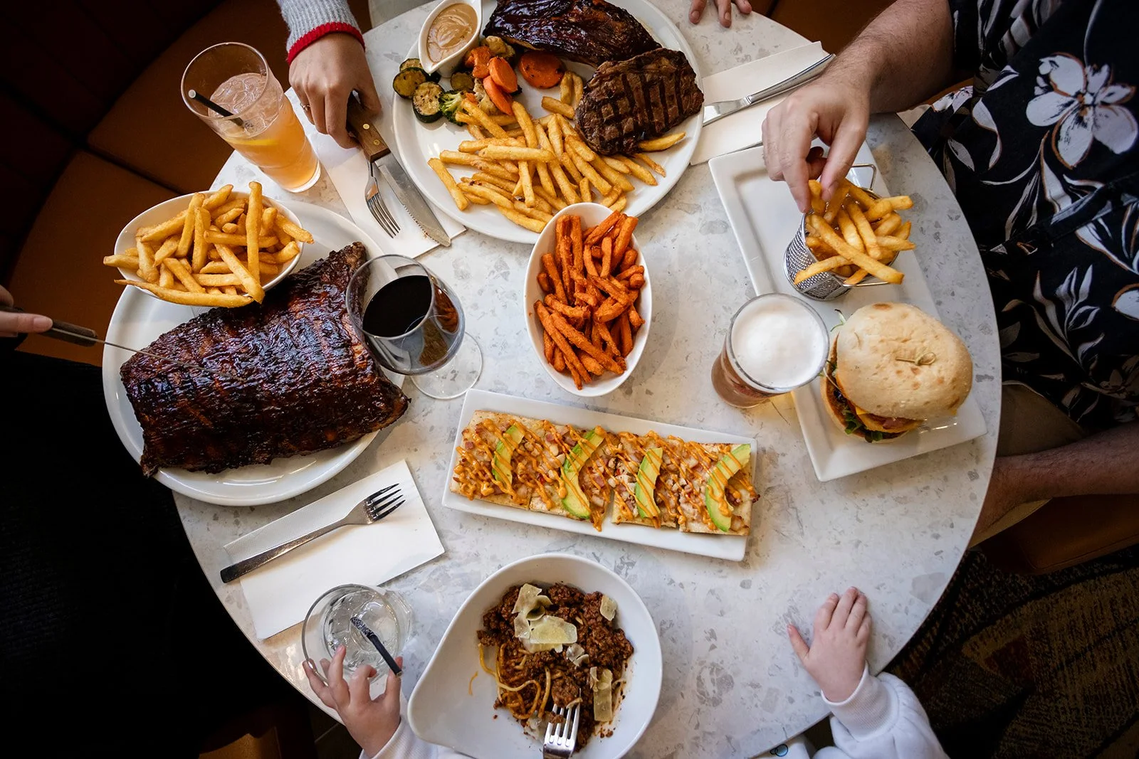 A table filled with various foods including French fries, a large piece of smoked ribs, grilled meats, a burger, a bowl of chili, a small pizza, sweet potato fries, and drinks.