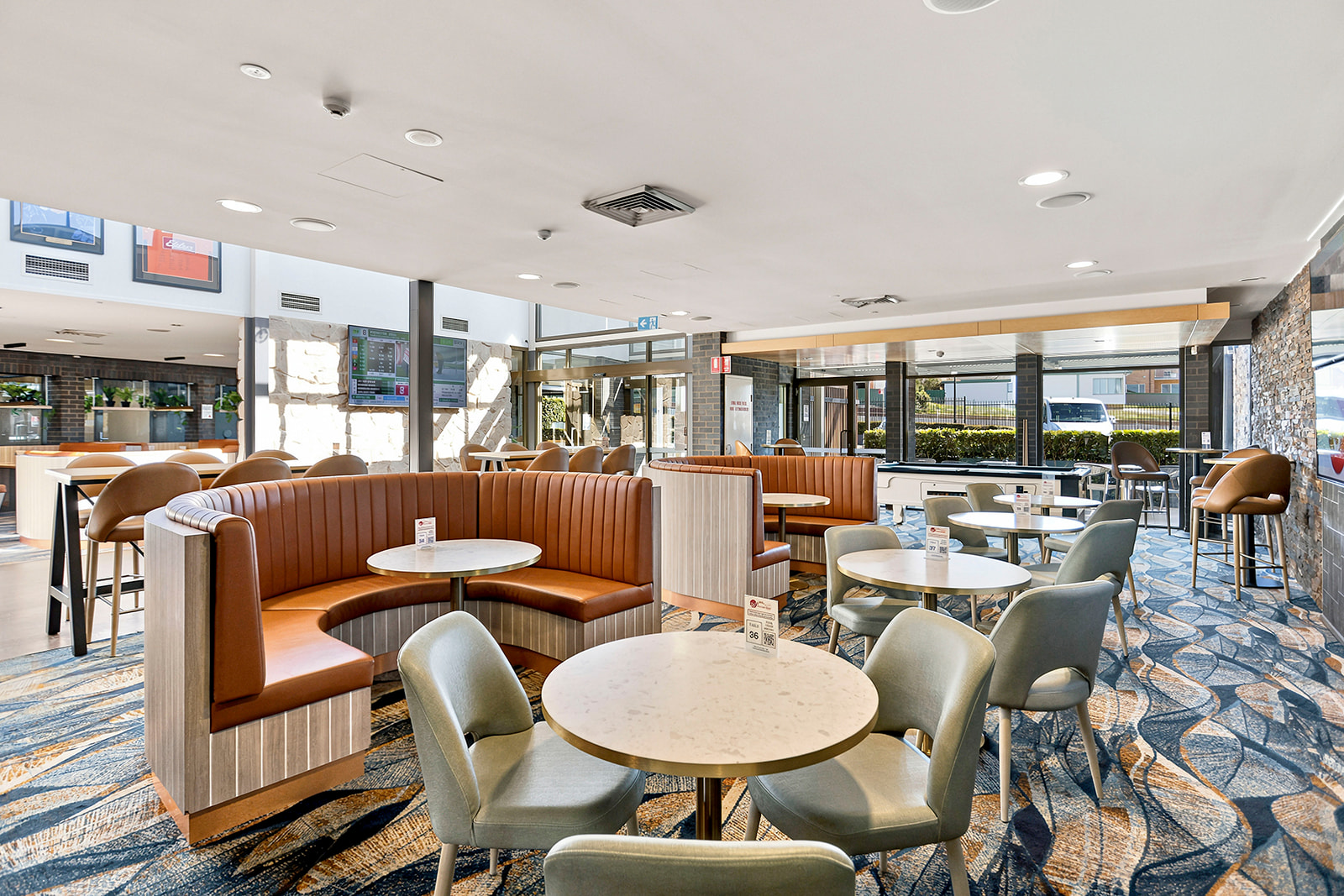 A modern, well-lit cafe with round tables, cushioned chairs, and booth seating, featuring a patterned carpet, exposed brick and stone walls, and large windows with outdoor views.
