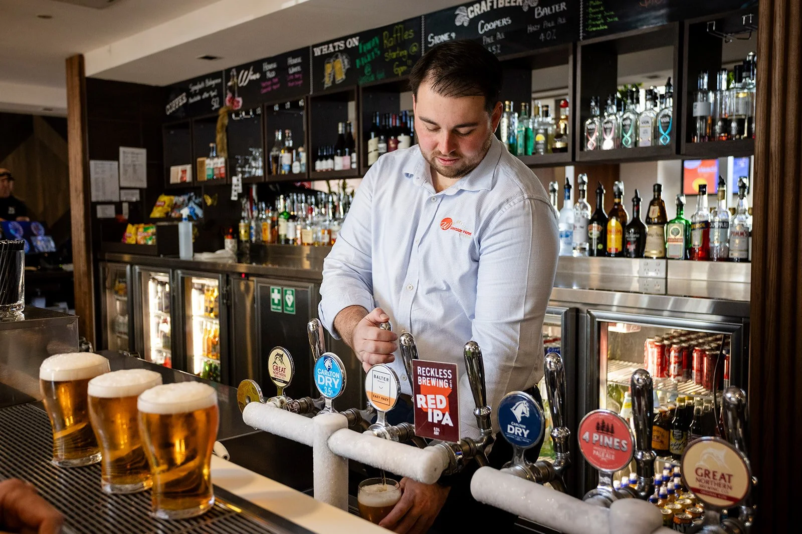 A bartender pours beer from a tap at a bar, with multiple beer taps in front, behind him a bar with bottles of liquor and a menu board above.