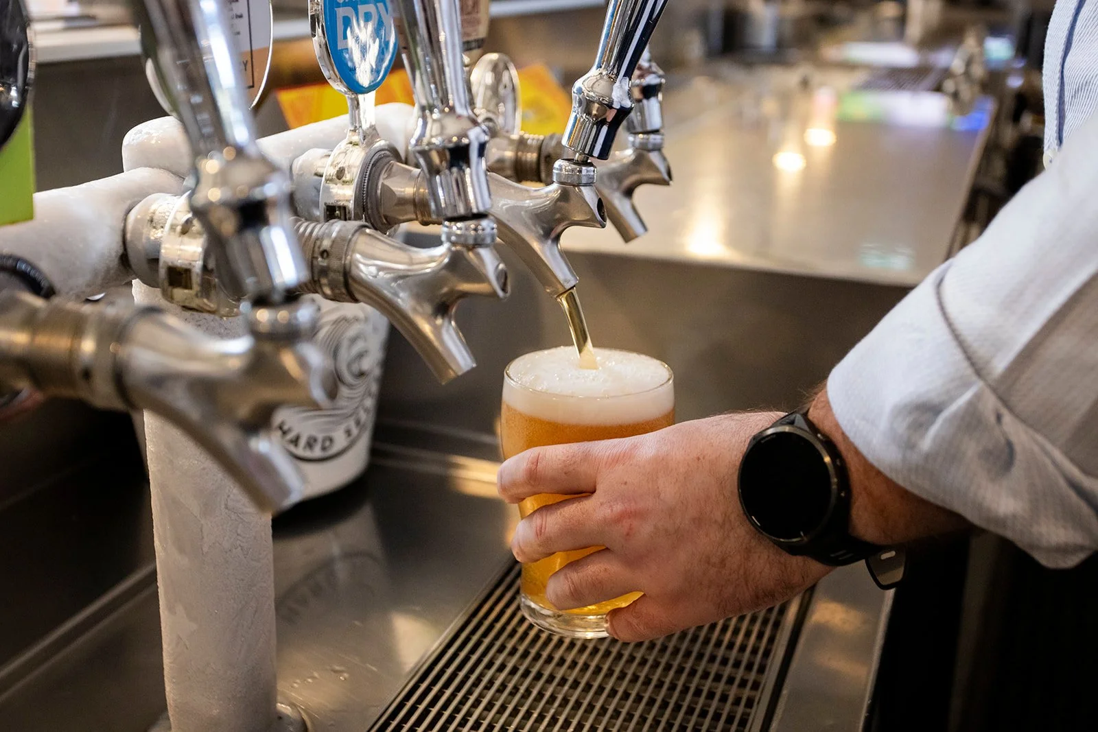 A person wearing a watch pours beer from a tap into a glass at a bar or brewery.