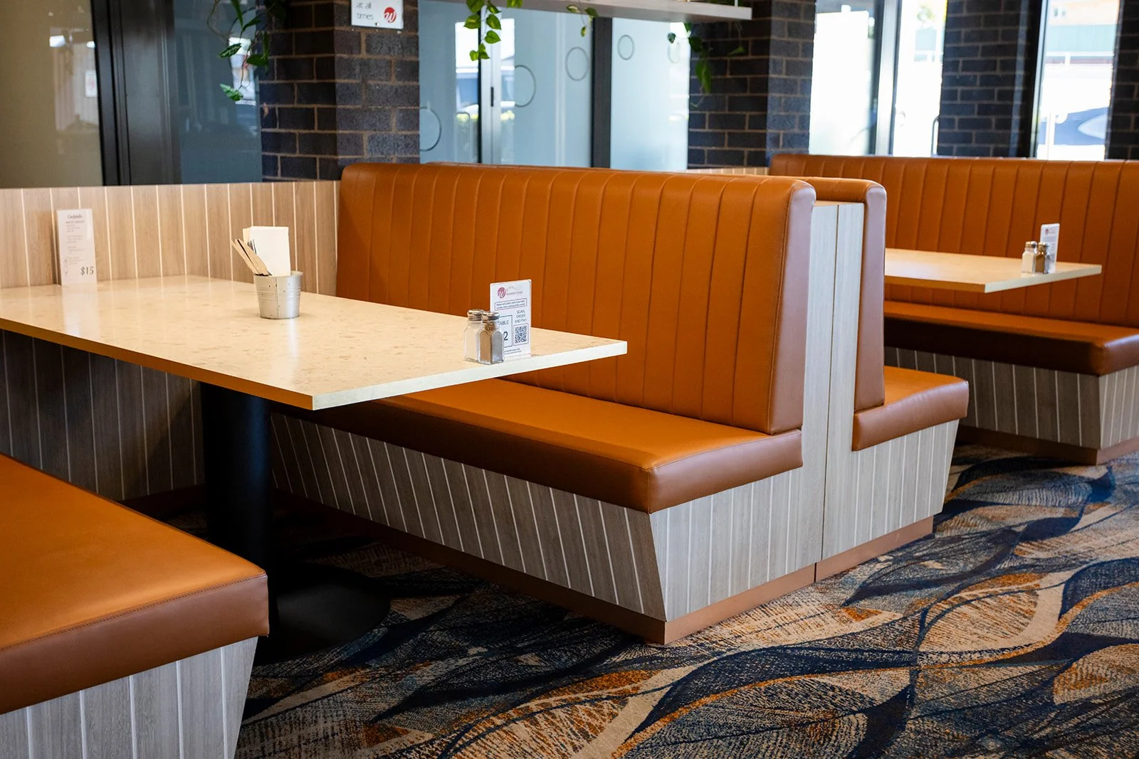 Empty restaurant booth seating with orange leather upholstery, light wood paneling, a white marble table, salt and pepper shakers, a small menu, and a window with brick wall and plants in the background.