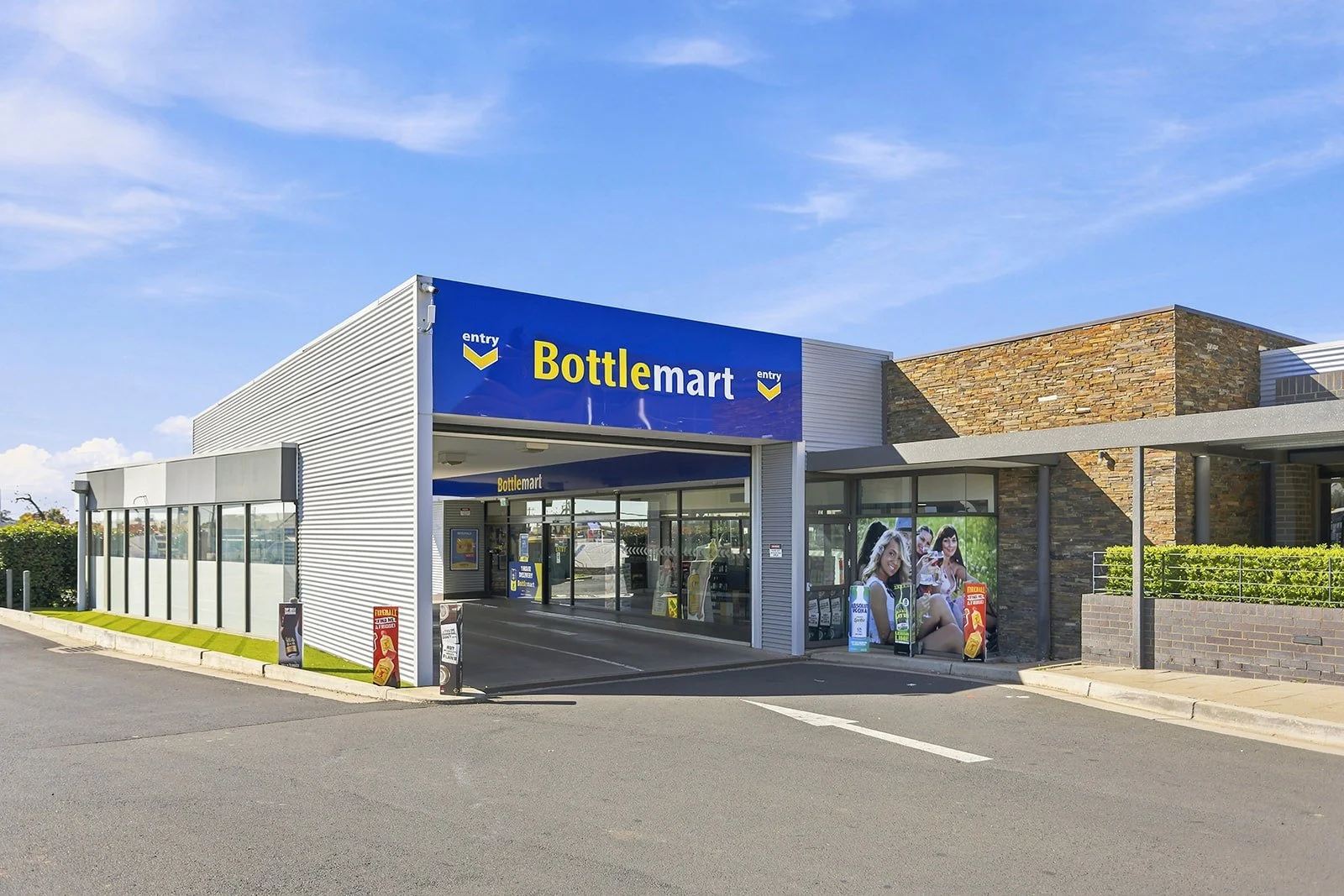 A Bottlemart liquor store with an entry drive-thru, featuring large glass windows, promotional banners, and a modern exterior with stone and metal siding, under a blue sky.
