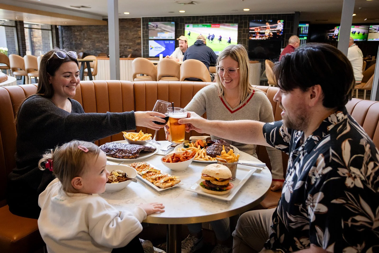 A group of five people, including a young girl, are gathered around a table in a restaurant, enjoying a meal and drinks, with a background of sports on TV screens.