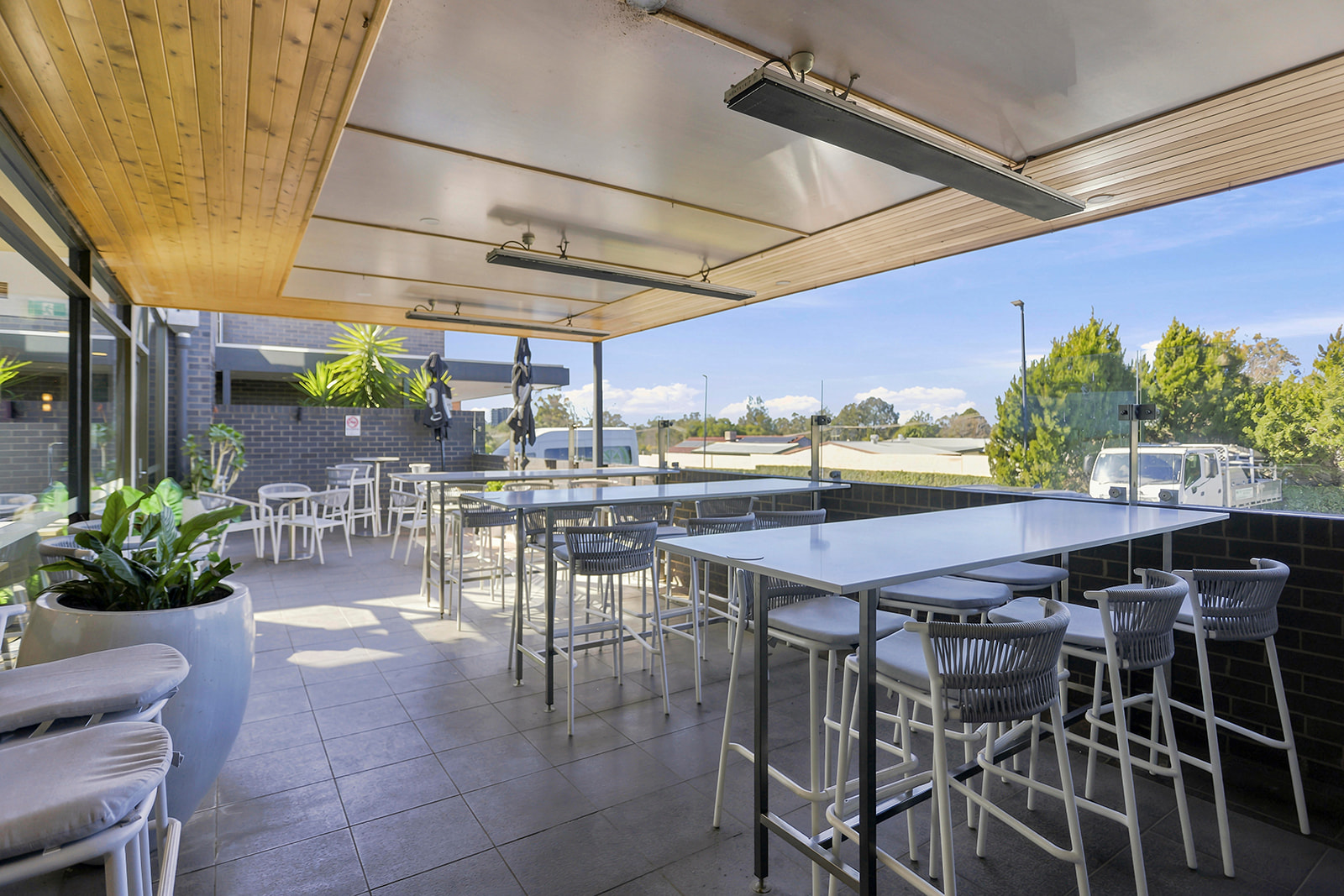 Outdoor patio with white tables and chairs, potted plants, glass railing, and a view of trees and a blue sky.