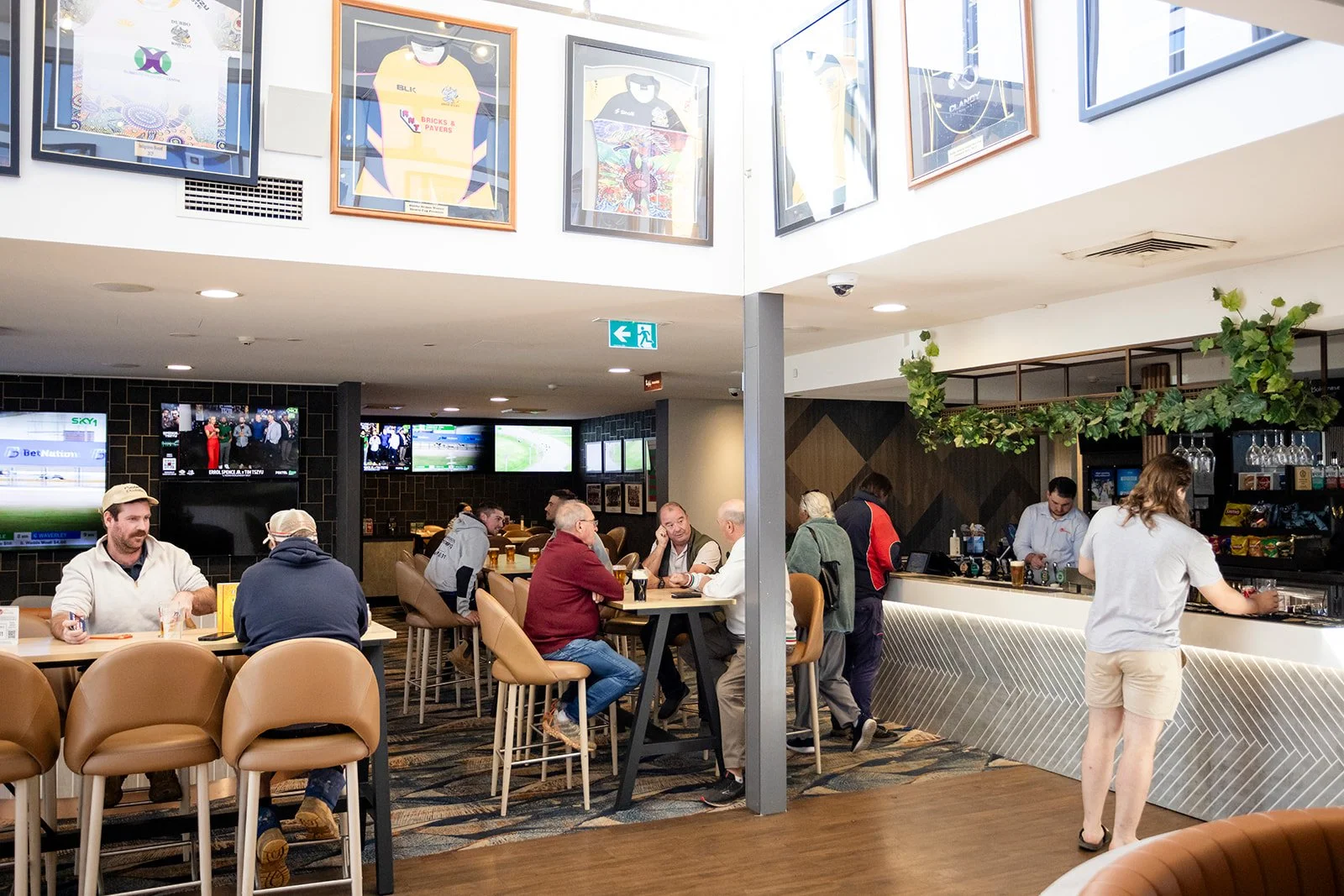 People sitting and standing in a bar or restaurant with multiple TVs on the wall, a barista or bartender preparing drinks, framed sports jerseys hanging above, and a woman with long hair and shorts near the bar.
