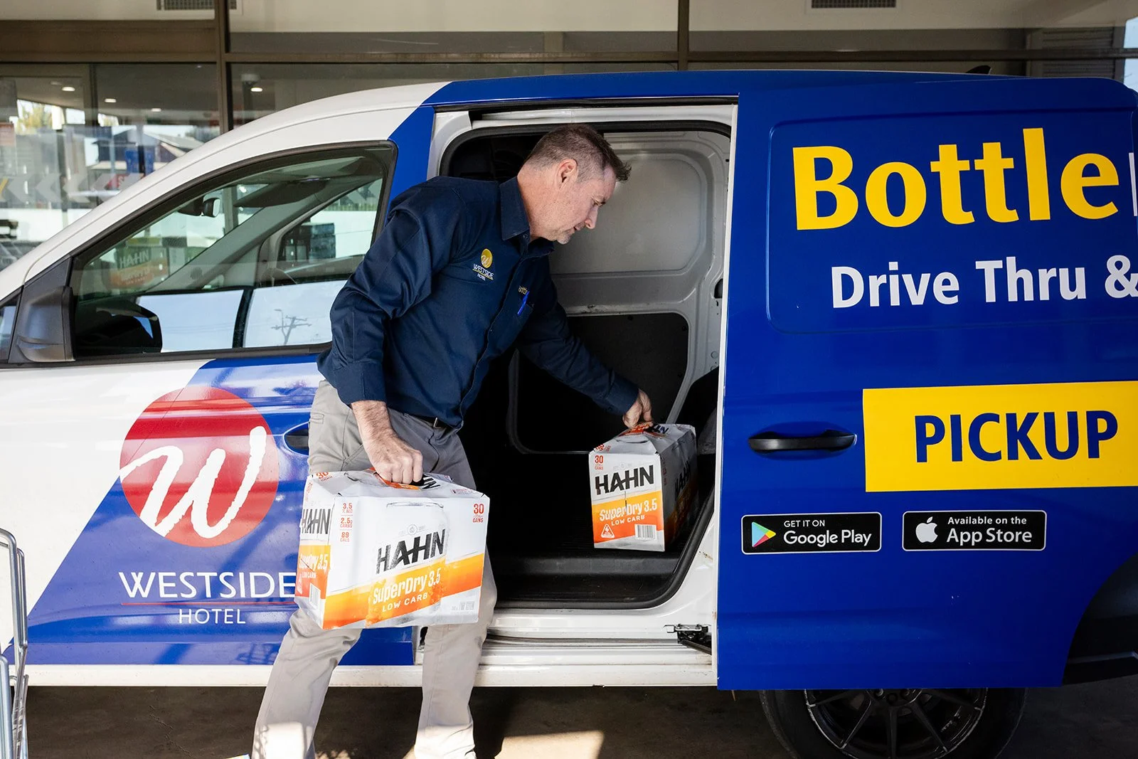A man loading boxes of Hahn SuperDry low carb beer into a pickup truck at a drive-thru pickup location, with branding of a hotel and app download icons visible.