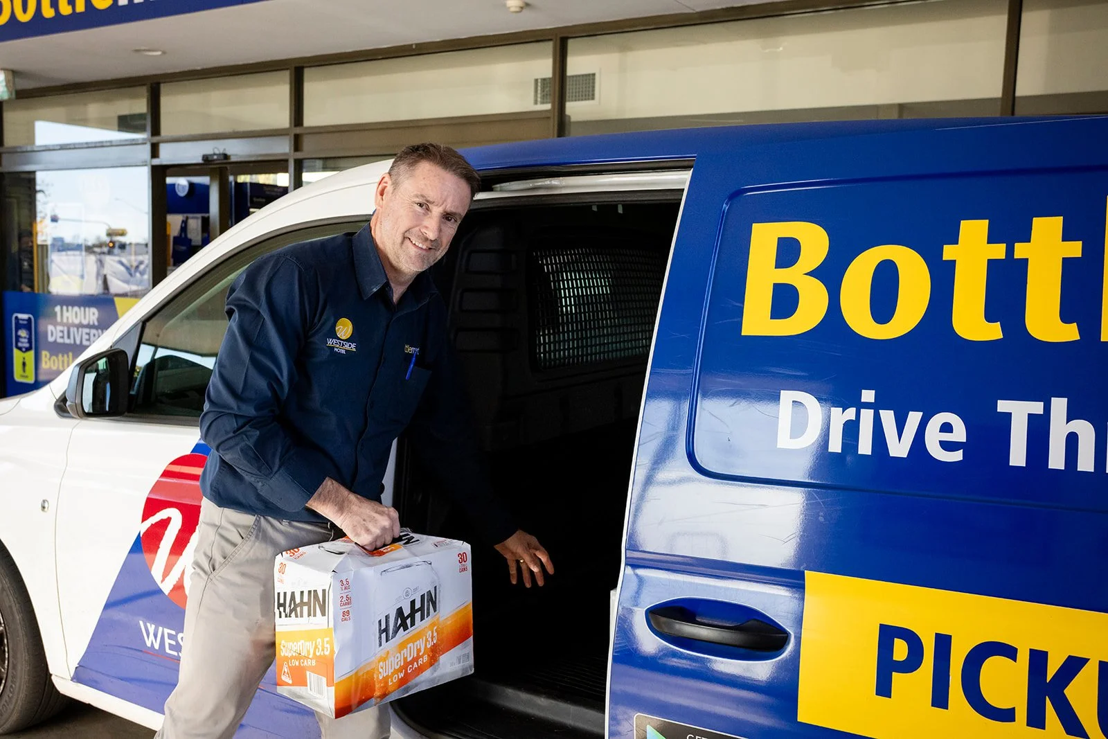 A man loading a box labeled HAMIN SuperDry 3.5 Low Carb into a blue Westside Pickup Drive Thru van.