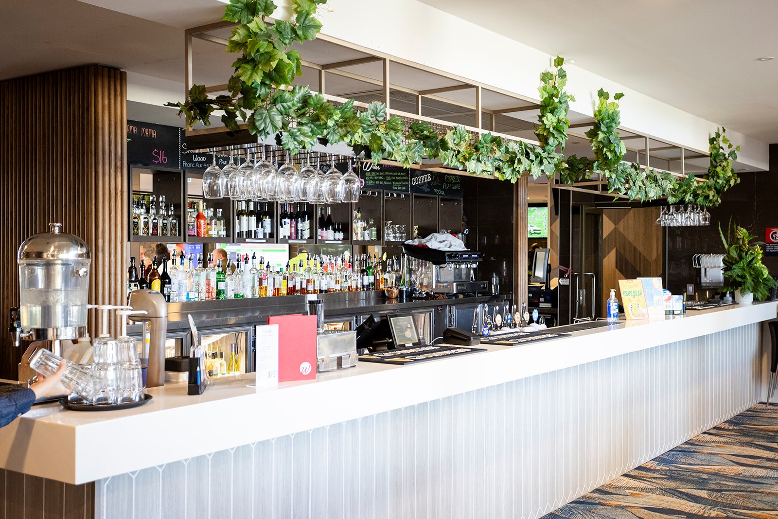 A modern bar with a white counter and a variety of bottles of alcohol displayed behind it. Hanging wine glasses are suspended above the counter, and green decorative plants are draped over a wooden frame above the bar. There is a hand reaching for a glass near a beverage dispenser on the counter.
