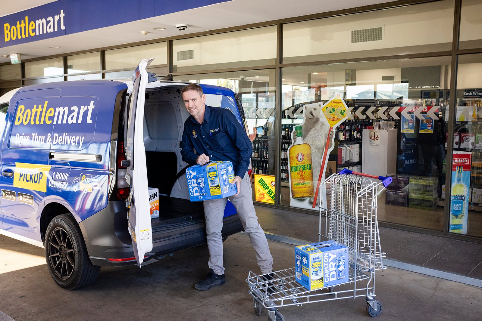 A man unloading cartons of Carlton Dry beer from a BottleMart delivery van at a store entrance. The store behind has large windows with shelves of products inside, and promotional posters, including one with a bear holding a bottle of Drop Bear Pineapple cider.