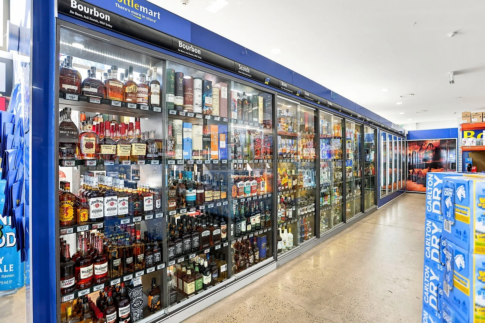 A grocery store aisle with a large refrigerated section filled with bottles of whiskey and other spirits. To the right, there are cases of Carlton Dry beer. The store has a blue and white color scheme with the store logo at the top.