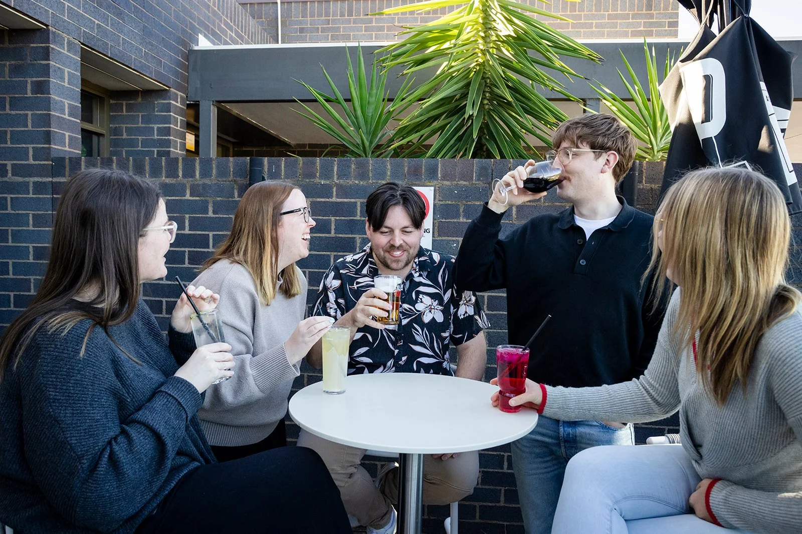Group of five friends enjoying drinks at an outdoor gathering, laughing and chatting around a small round table, with a brick wall and large green plants in the background.