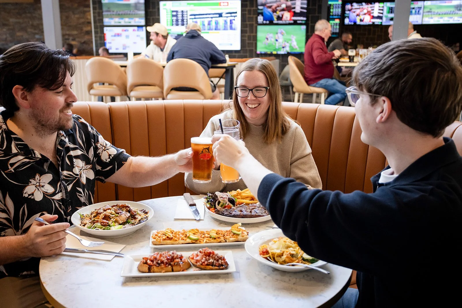 Three friends clinking glasses of beer and enjoying a meal together at a restaurant, with shared dishes of pizza, pasta, and salads on the table, while others are watching sports on multiple TV screens in the background.
