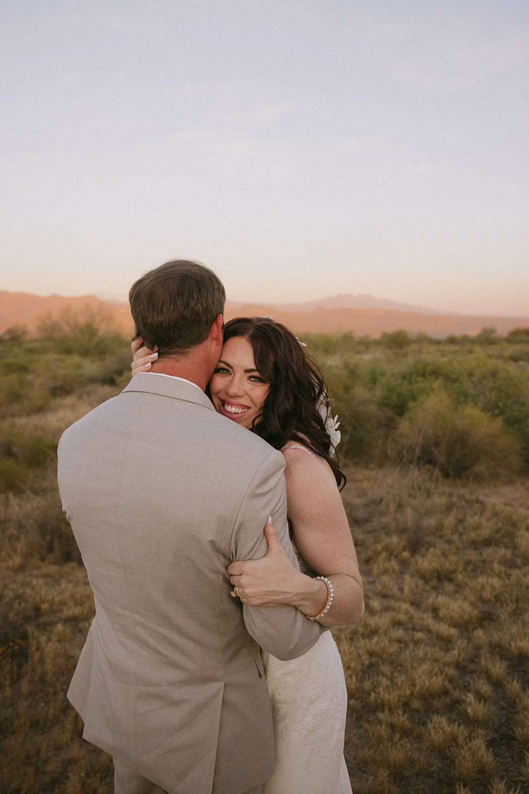 A couple in wedding attire embracing outdoors during sunset, with desert landscape and mountains in the background.