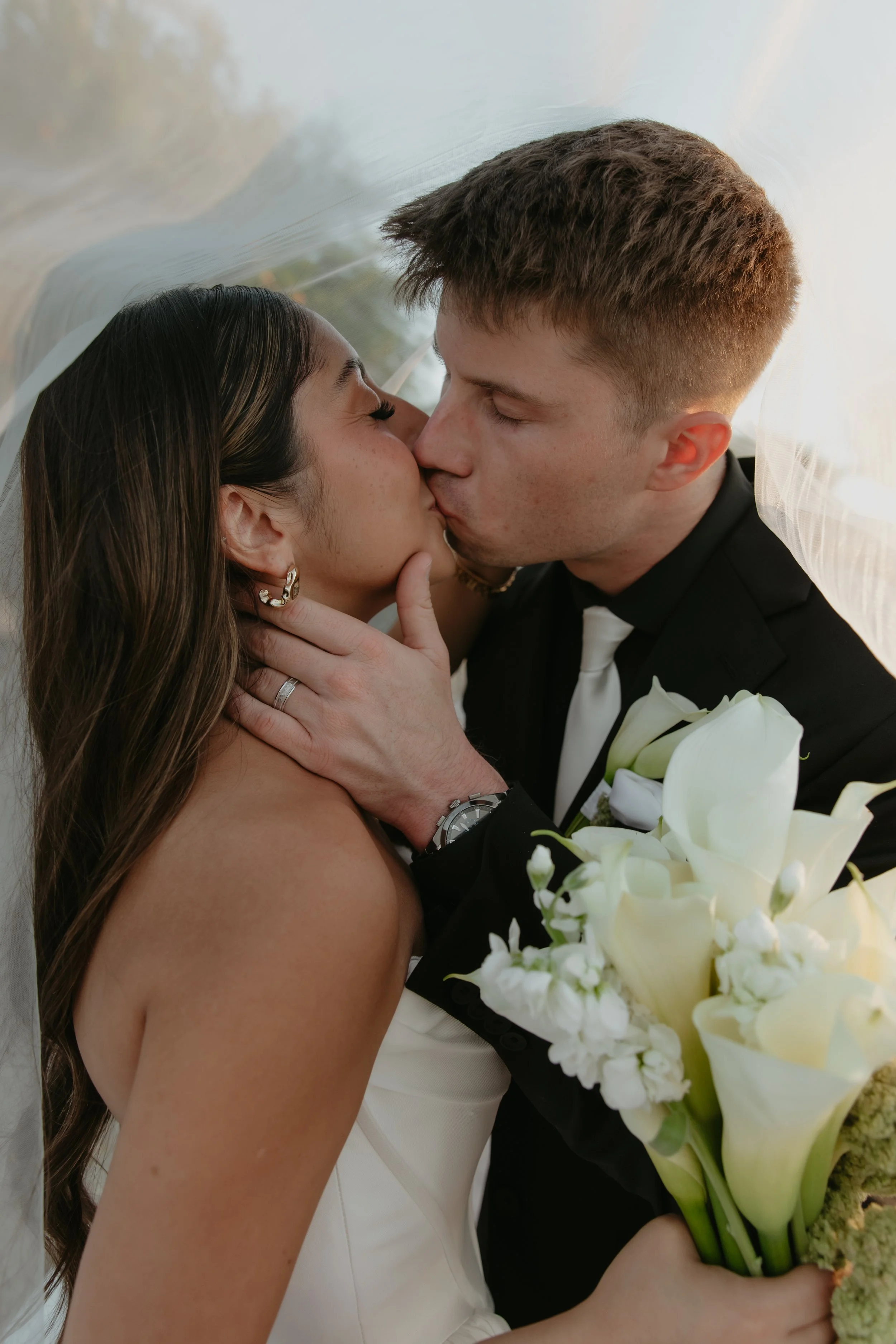 A bride and groom sharing a kiss, with the bride holding a bouquet of white calla lilies and other white flowers, under a wedding veil. The bride has long dark hair and is wearing earrings; the groom has short light brown hair and is dressed in a tuxedo.