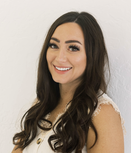 A young woman with long dark hair and makeup, smiling and looking at the camera, wearing a white sleeveless top, standing against a white wall.