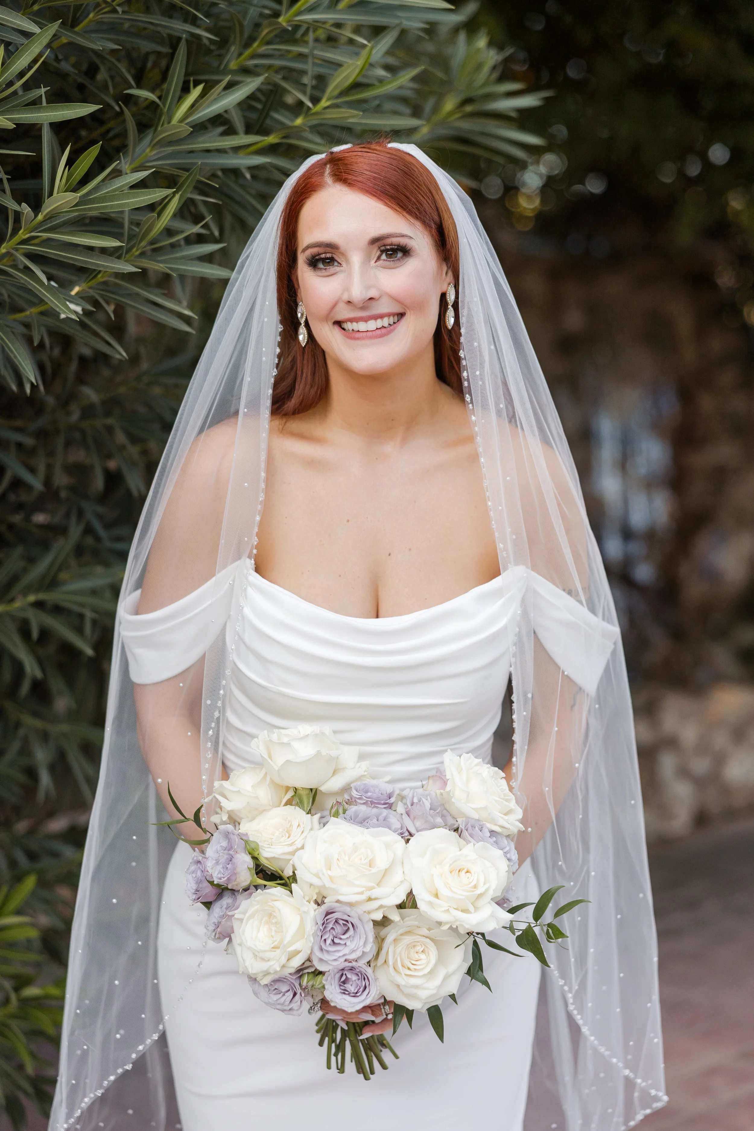 A bride with red hair wearing a white wedding dress and a veil, holding a bouquet of white and lavender roses, standing outdoors with green foliage in the background.