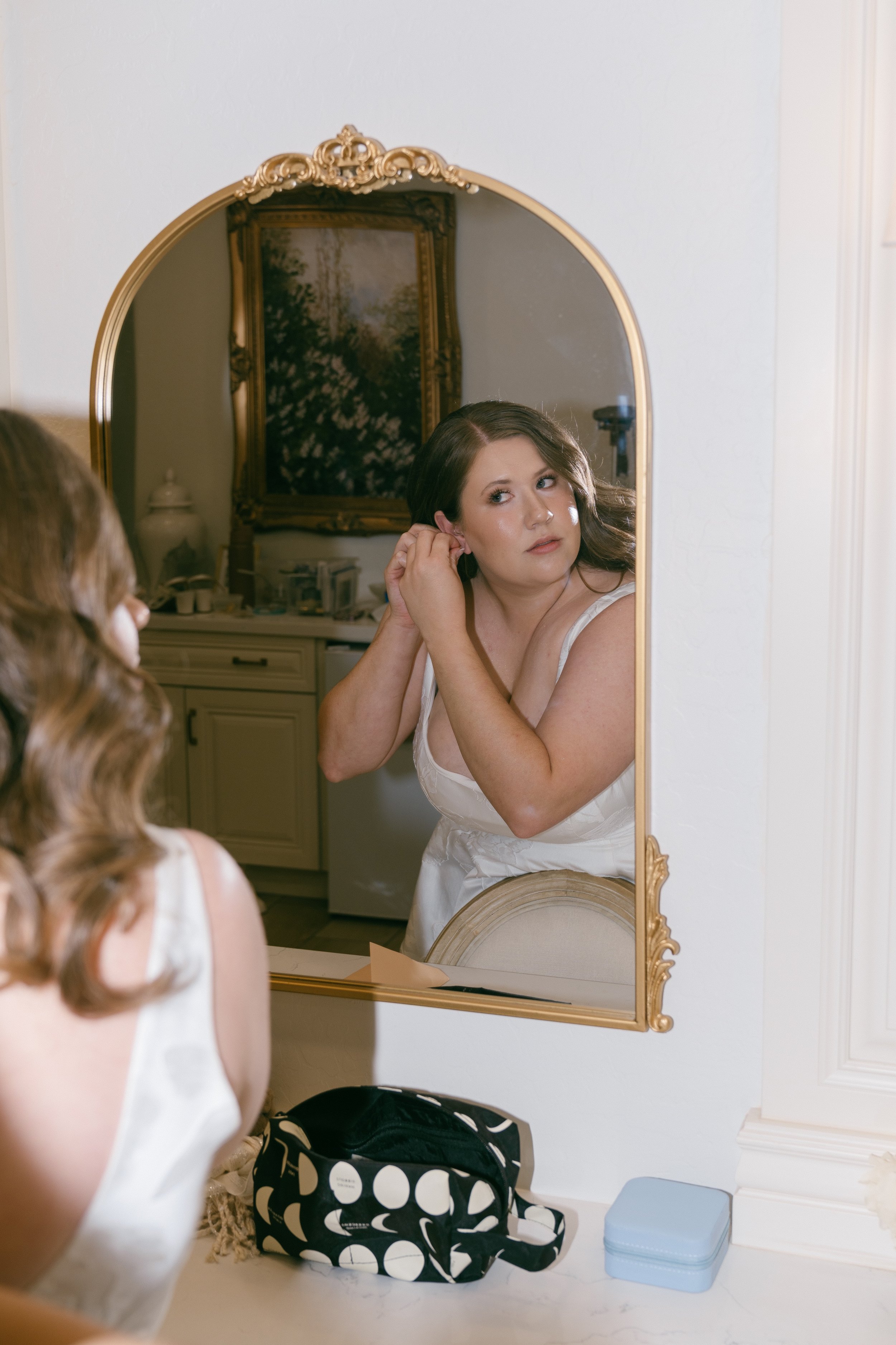 Woman with brown hair looking into mirror, adjusting earring, wearing white dress, in a room with a decorative gold-framed mirror and dresser, with a black and white striped bag and blue jewelry box in the foreground.