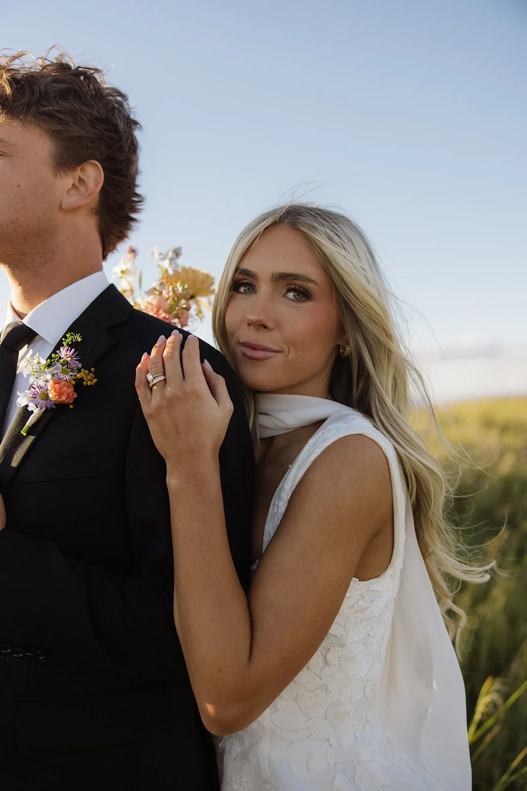 A woman with blonde hair, wearing a white dress, gently resting her hand on a man's shoulder, who is dressed in a black suit with a floral boutonniere. The woman is looking at the camera with a soft expression, and there is a background of open sky and greenery.