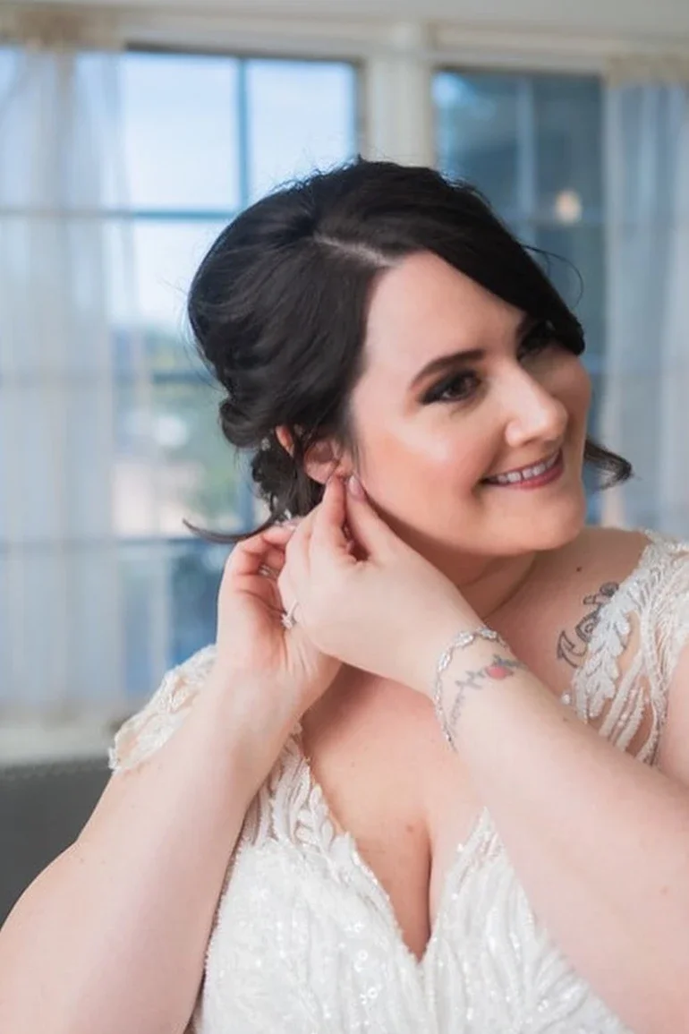 Smiling woman with dark hair and tattoos adjusting her earring in a bright room with large windows.