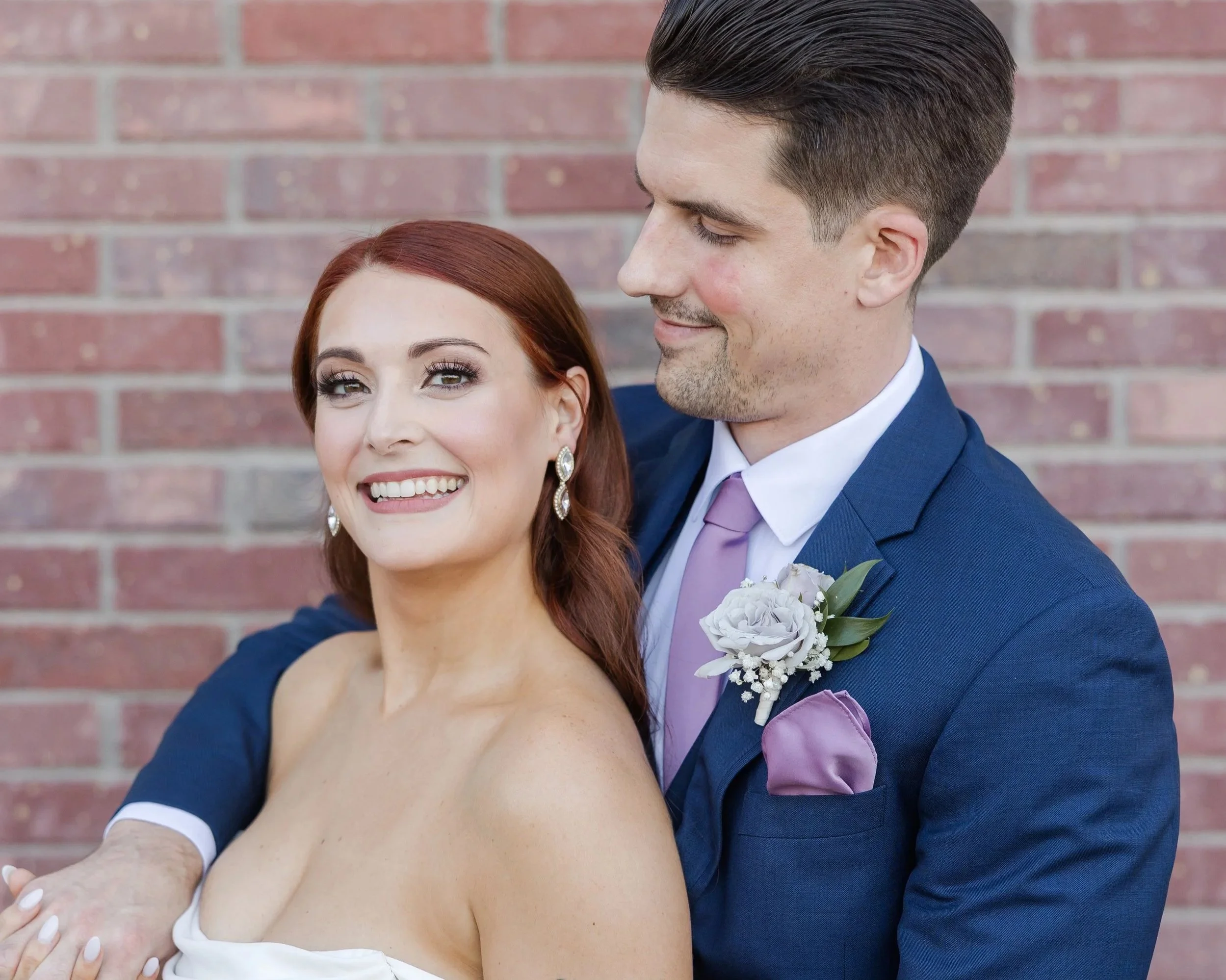 A happy couple dressed in formal attire, standing close together against a brick wall. The woman has red hair and is smiling, while the man has dark hair and is smiling with his eyes closed, wearing a navy blue suit, white shirt, and lavender tie, with a boutonniere on his lapel.