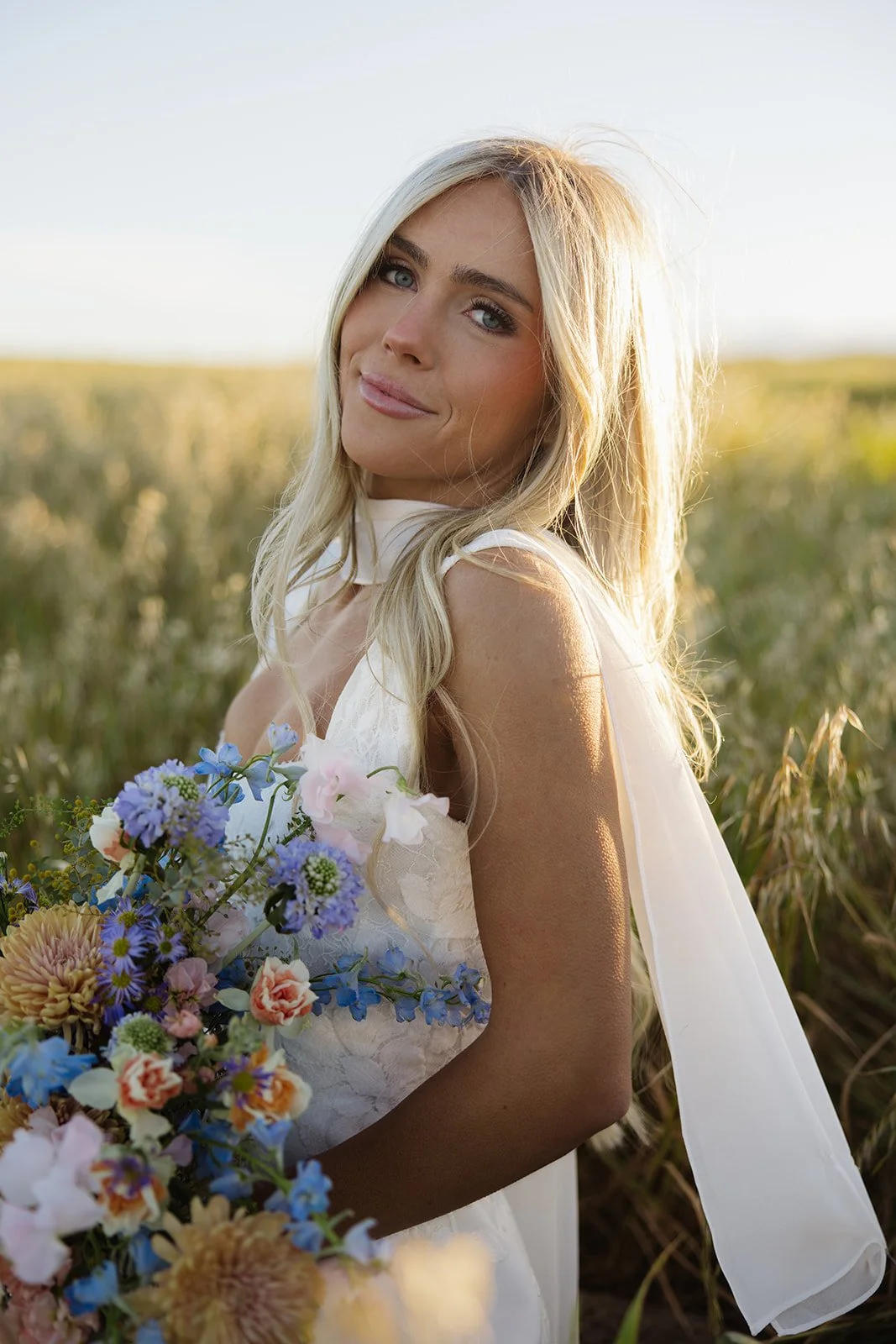 A woman with long blonde hair holding a large bouquet of colorful flowers, standing in a field of tall grass during sunset.