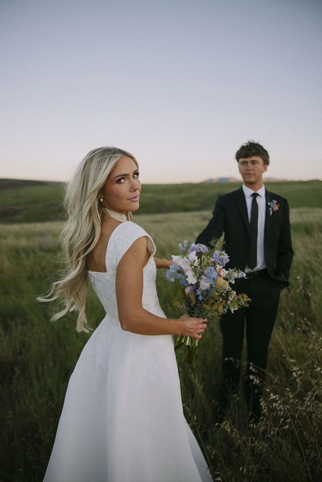 A bride in a white wedding dress holding a bouquet of flowers, with a groom in a black suit and tie in the background, standing in a grassy field during sunset.