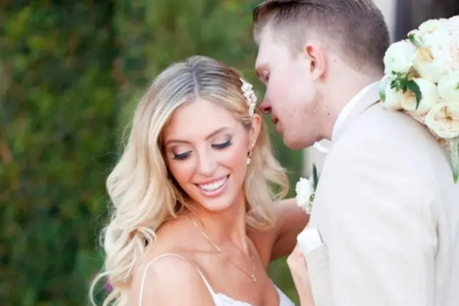 A bride and groom sharing a tender moment outdoors, with the bride smiling and the groom leaning in close, surrounded by greenery.