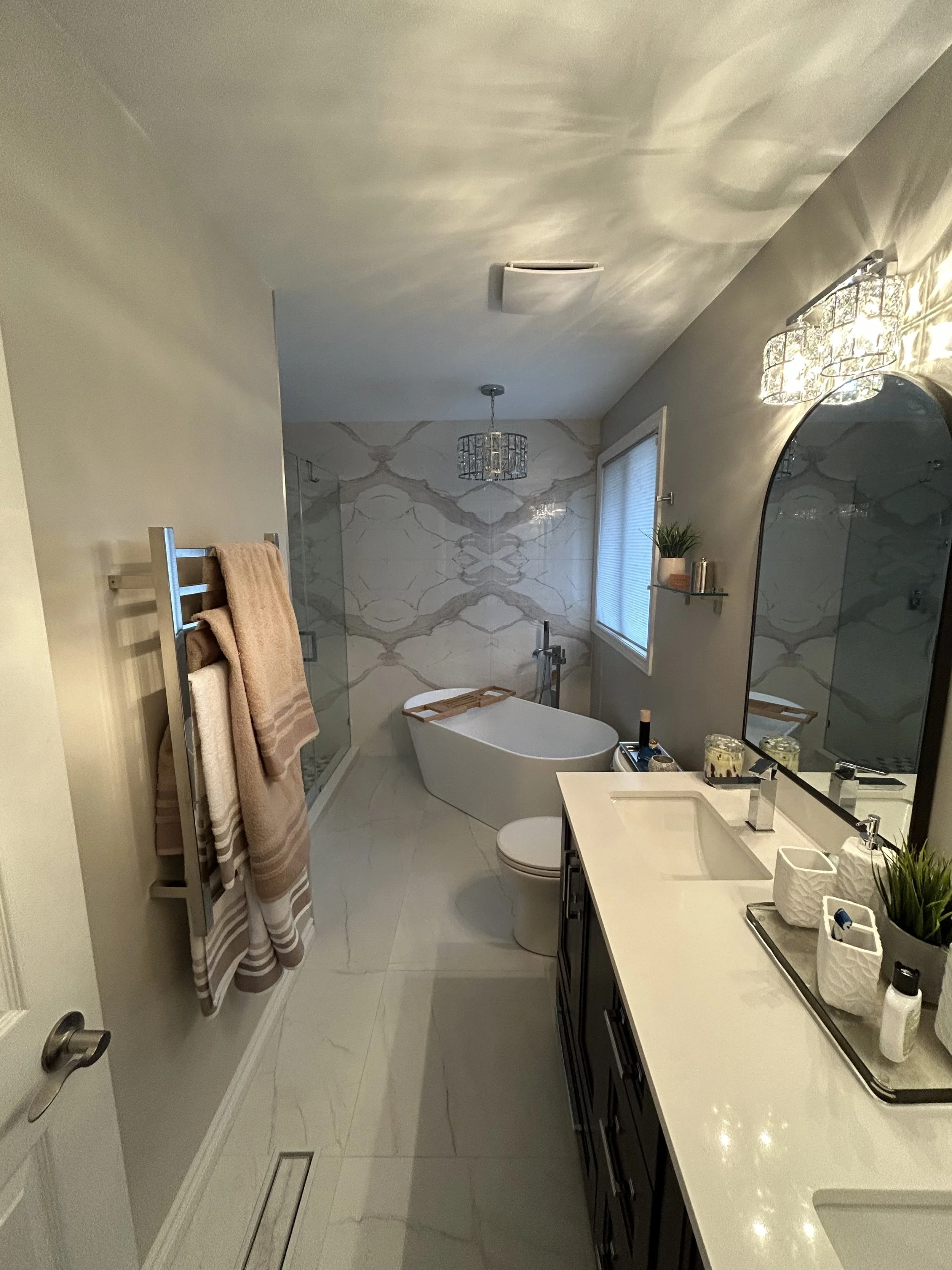A modern bathroom with a free-standing bathtub, a large mirror, dark vanity cabinets, and a window with blinds. There are towel racks with beige towels, and decorative candles on the countertop. The walls have a marble pattern, and light fixtures hang from the ceiling.