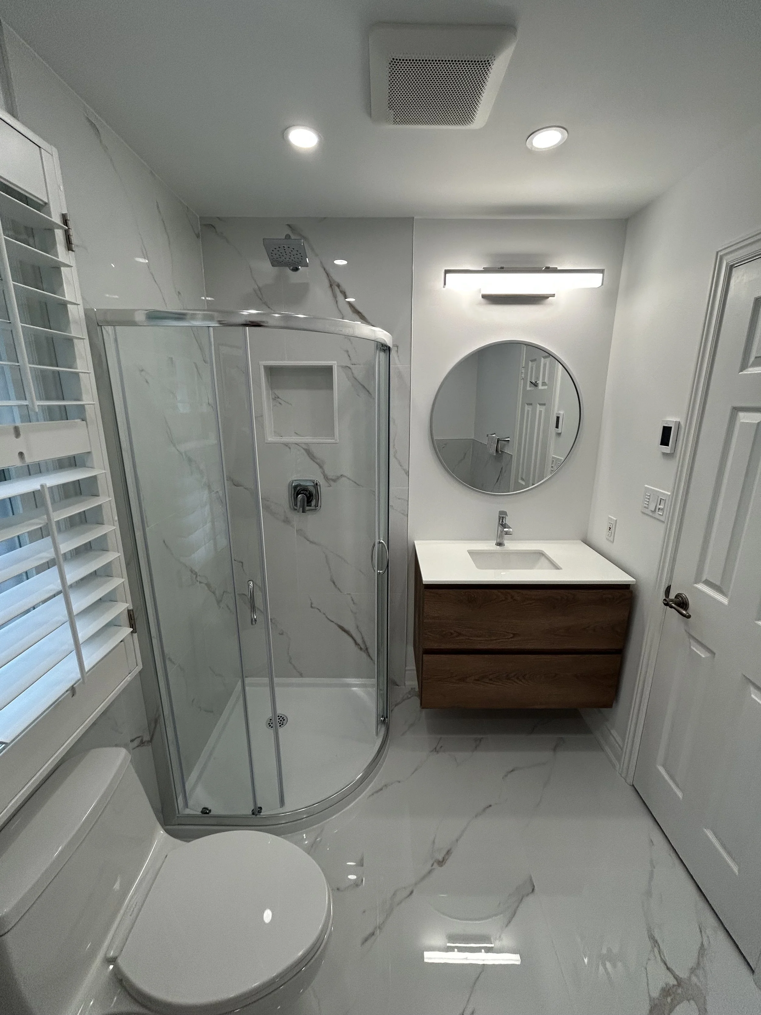 Modern bathroom featuring a glass shower enclosure with marble wall tiles, a white vanity with a round mirror and LED light above, a white toilet, and marble flooring.