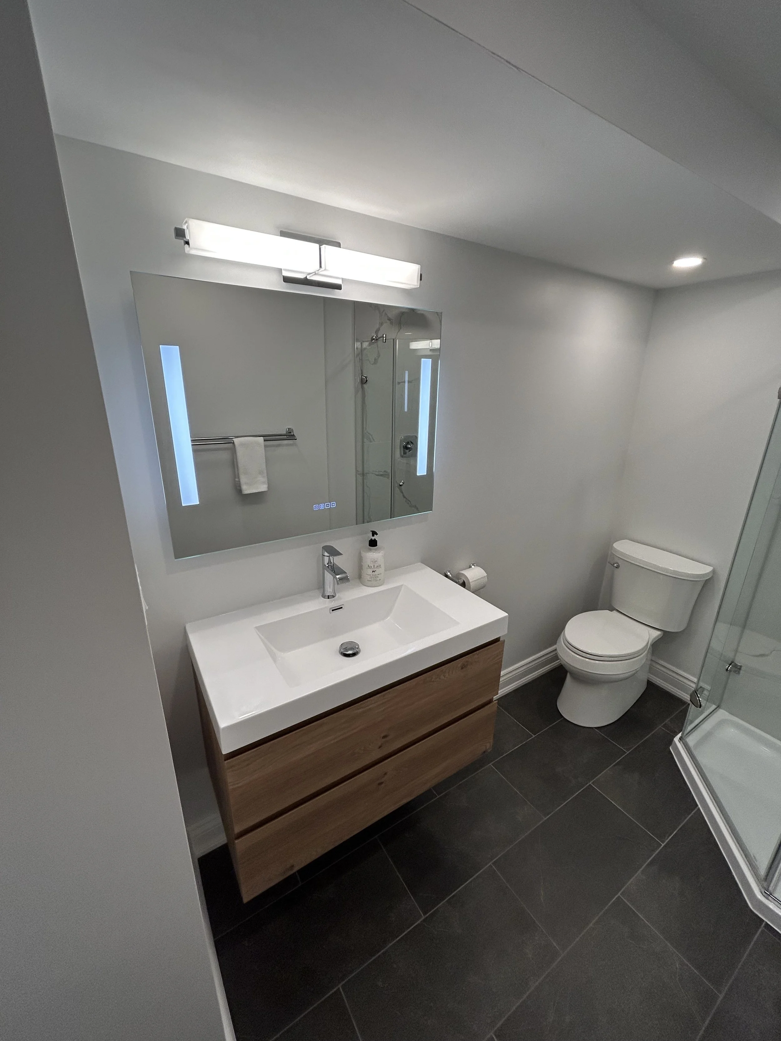 Modern bathroom with white walls, a rectangular mirror, a wall-mounted light fixture, a white sink with a wooden vanity, a soap dispenser, a toilet, and a glass-enclosed shower.