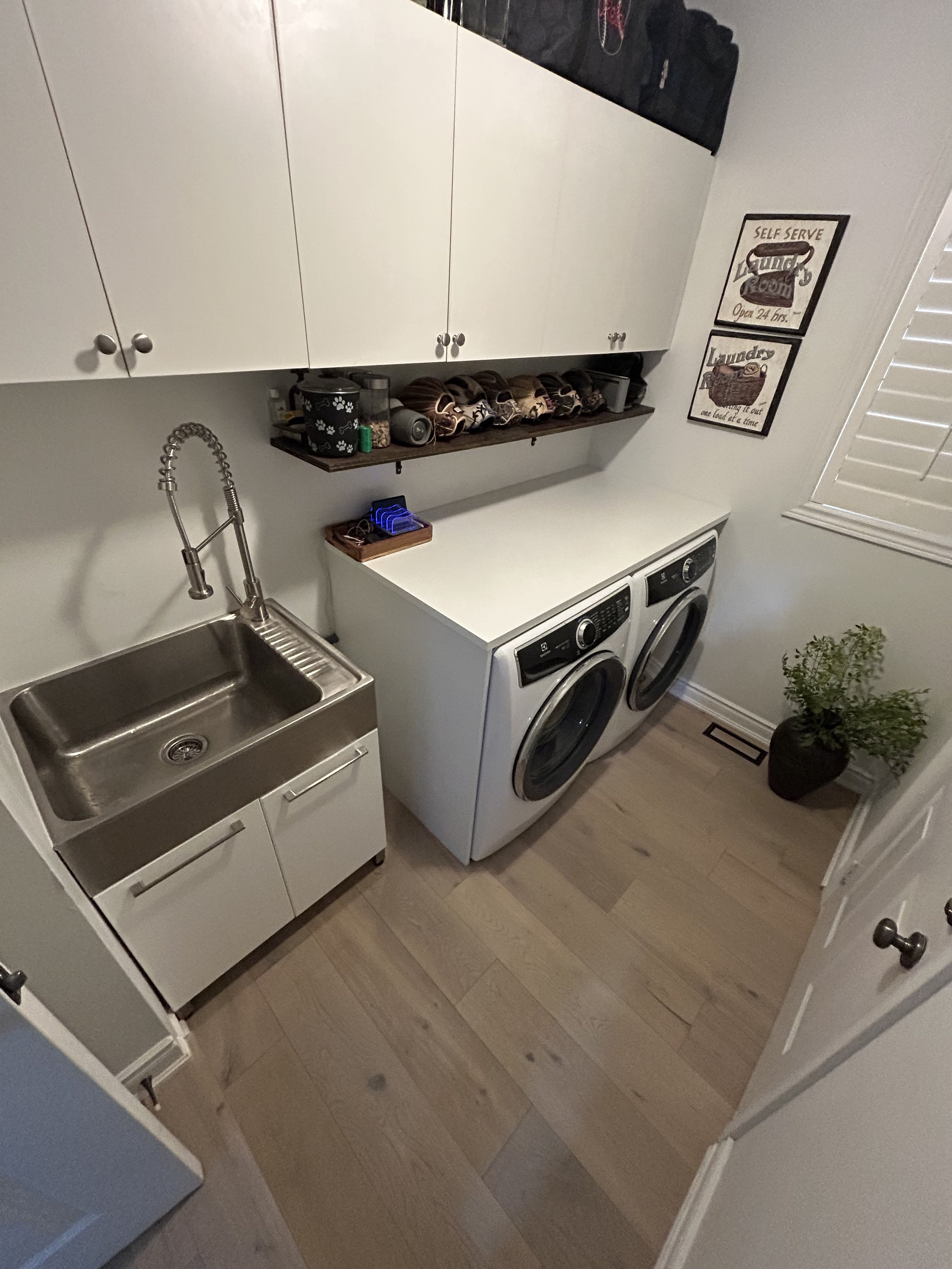 A laundry room with white cabinets, a stainless steel utility sink, a washing machine and dryer, a wooden shelf with shoes and small baskets, and framed wall art. There is a potted plant near the window with closed blinds.