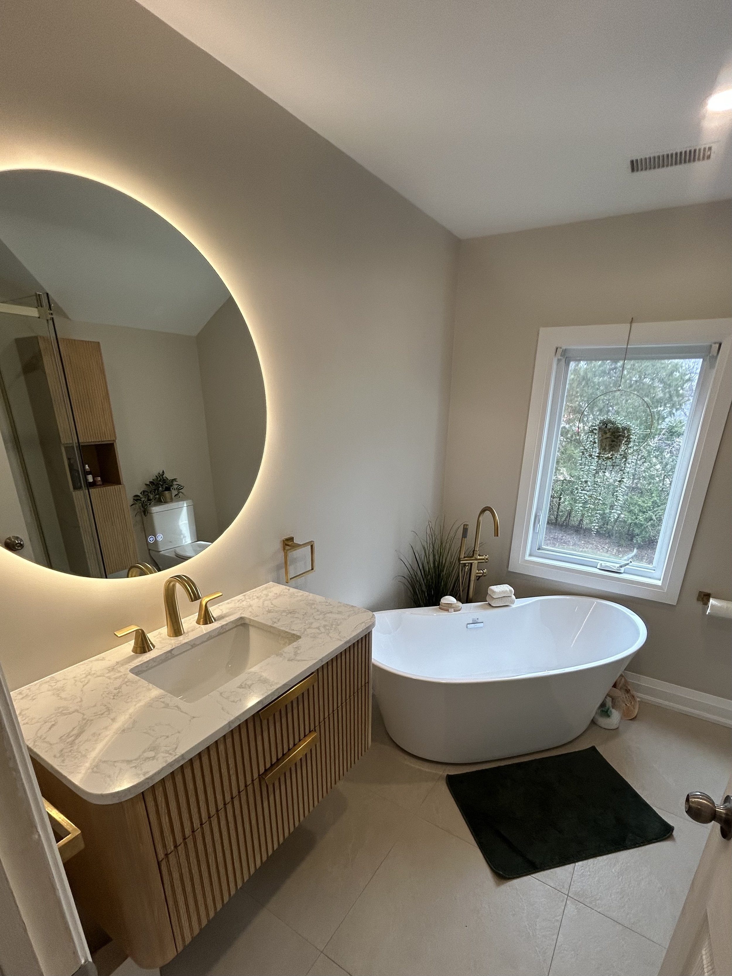 A bathroom with a large backlit round mirror above a marble countertop with two gold faucets. A white oval bathtub is near a window with trees outside. There are small towels on the bathtub rim, a plant in the background, and a black bath mat on the floor.