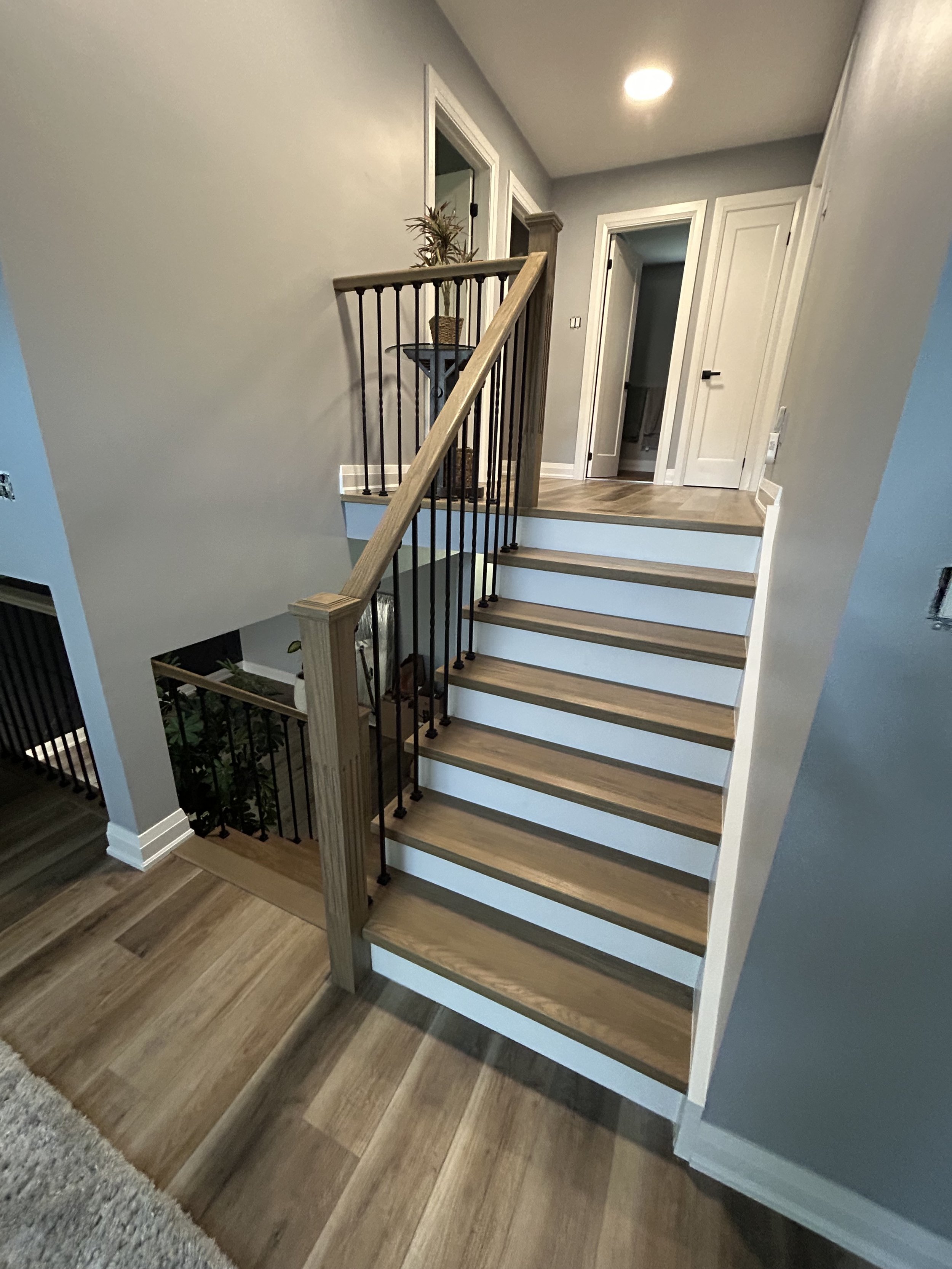 Interior view of a staircase in a modern home with wooden steps, a wooden handrail, and white risers, leading up to a hallway with doors and a gray wall.