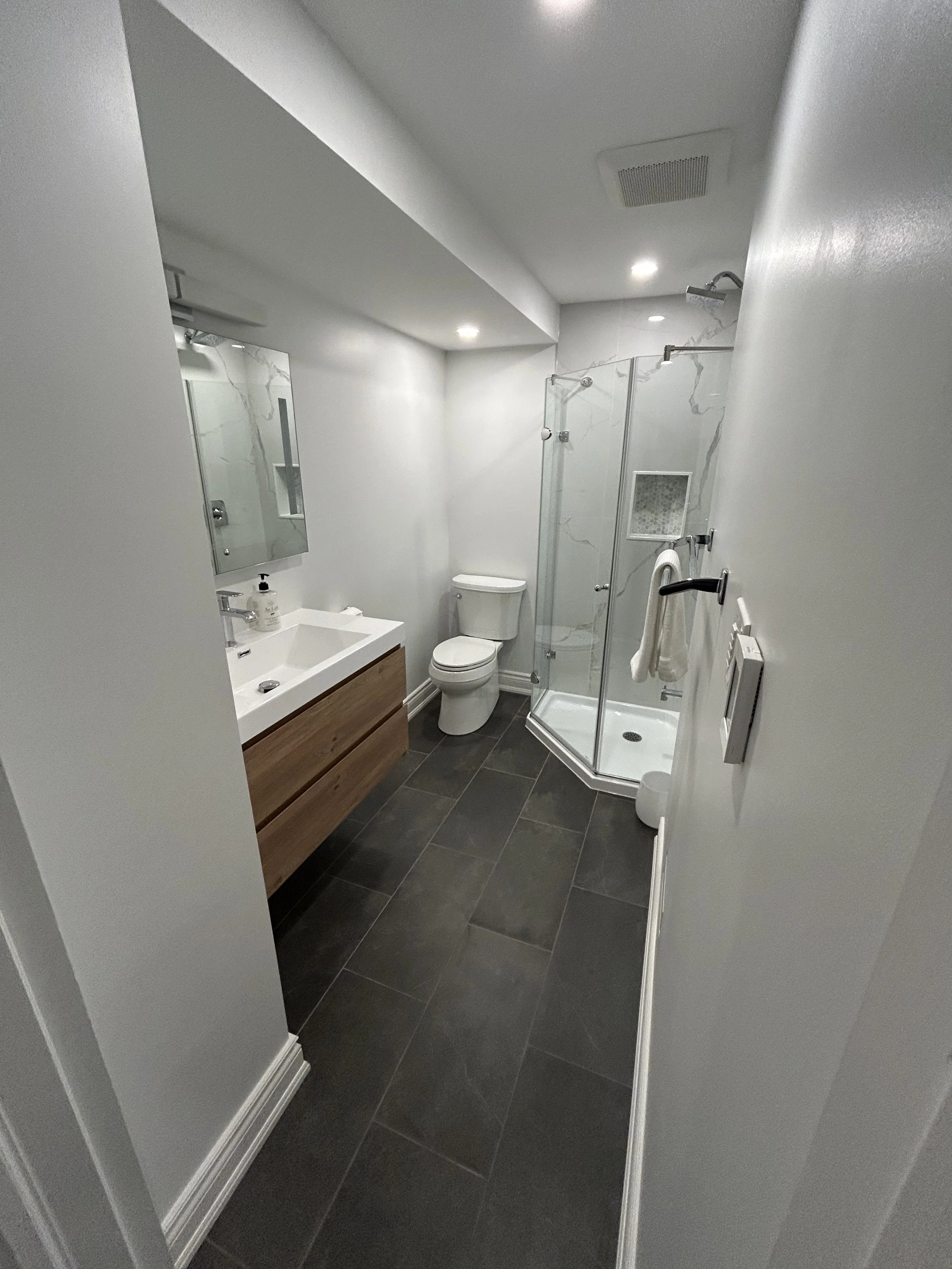 A modern bathroom with a white sink and wooden vanity, a white toilet, a small shower with glass door, a wall-mounted towel rack, dark tiled flooring, and white walls with marble accents.