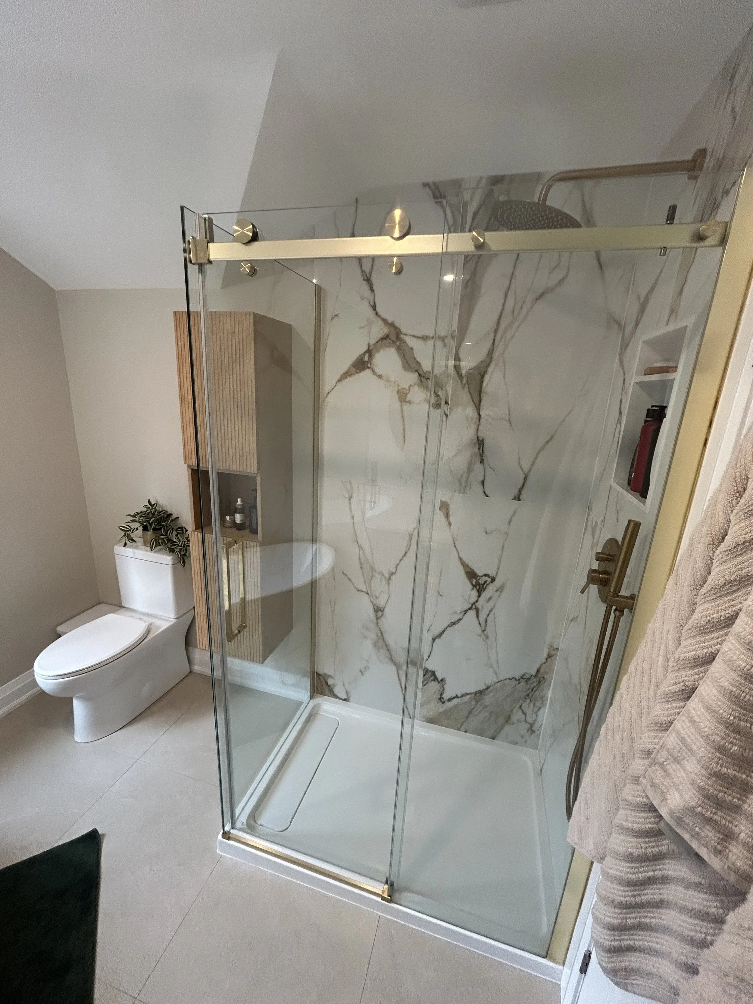 A bathroom with a toilet, a tiled shower with marble walls and glass door, a towel on a rack, and a plant on a wooden shelf.