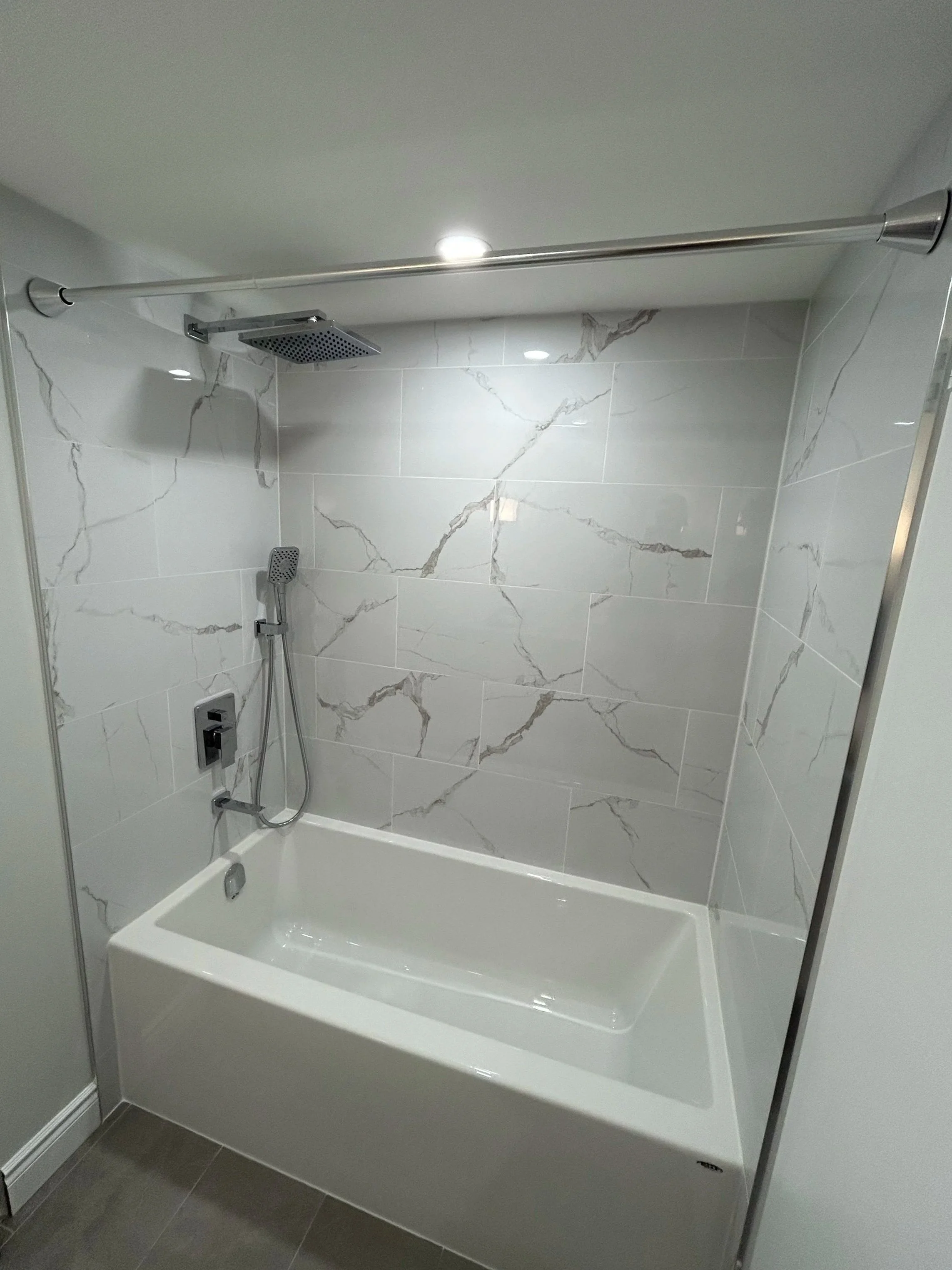 Bathroom shower with marble tile walls, a chrome shower head and a handheld shower wand, a square rain shower head, a faucet, and a white bathtub with a drain.