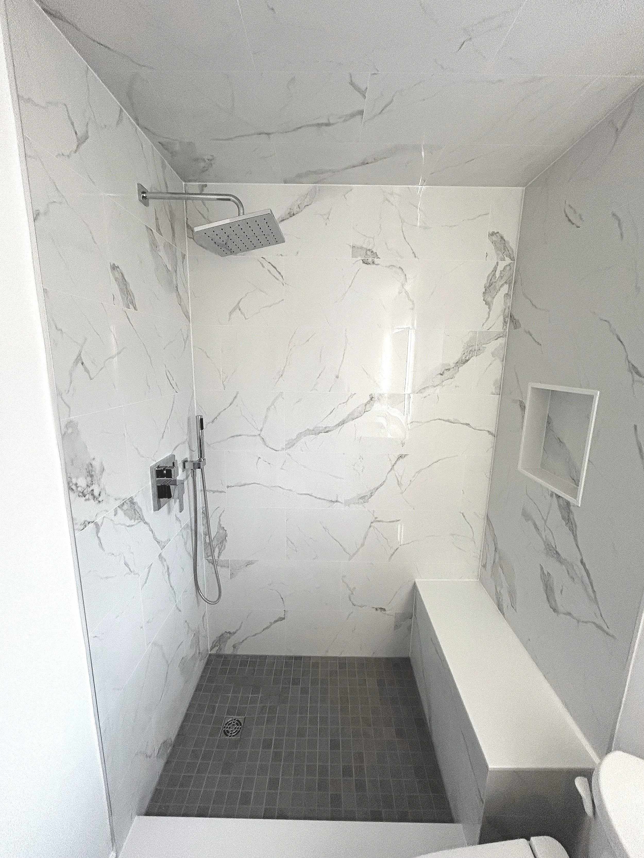A modern shower area with marble wall tiles, a rainfall showerhead, a handheld shower, a small built-in bench, and a tiled floor drain.