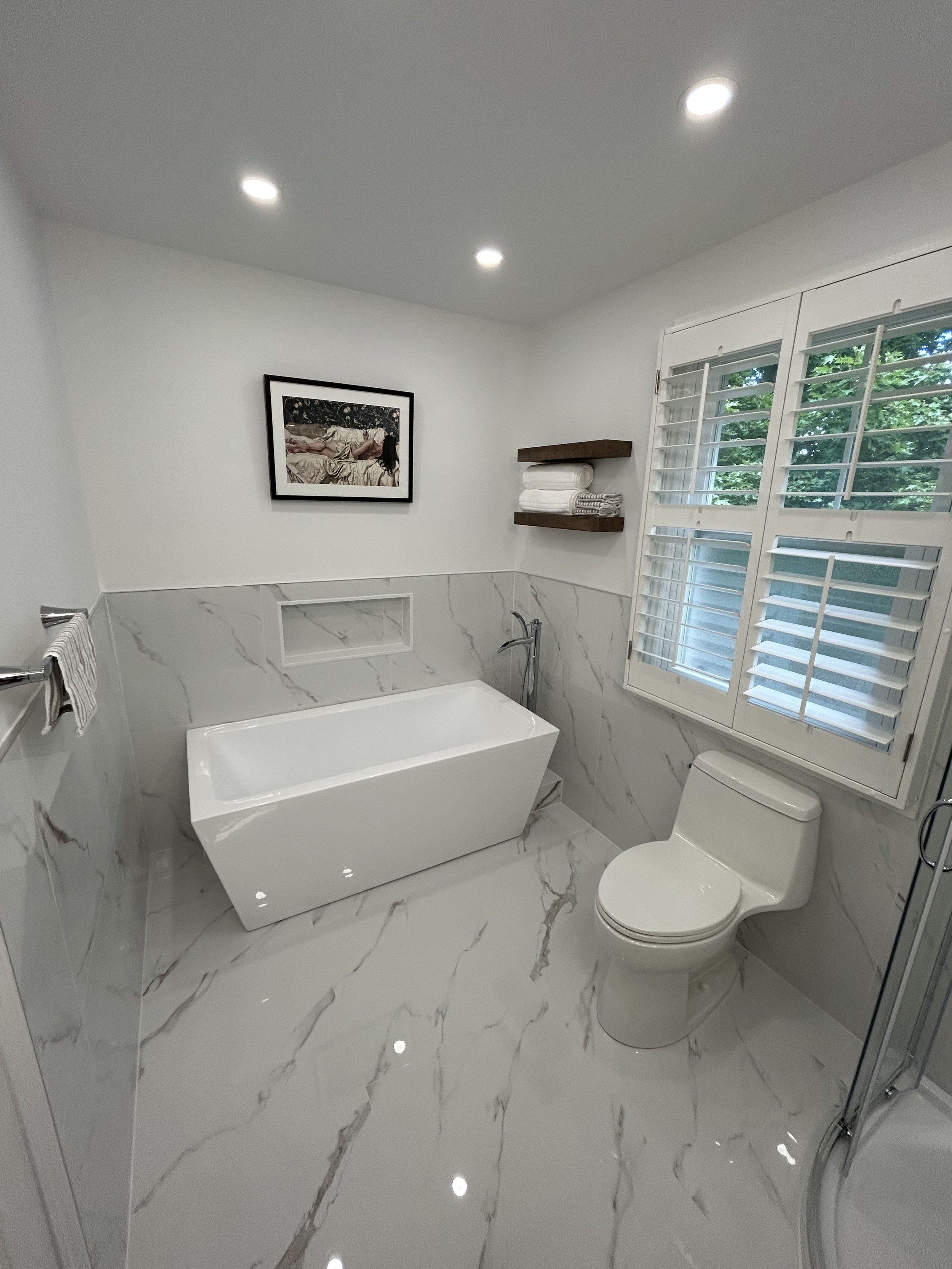 A modern bathroom with white marble walls and floor, a bathtub, a toilet, a window with white shutters, and a framed art piece on the wall.