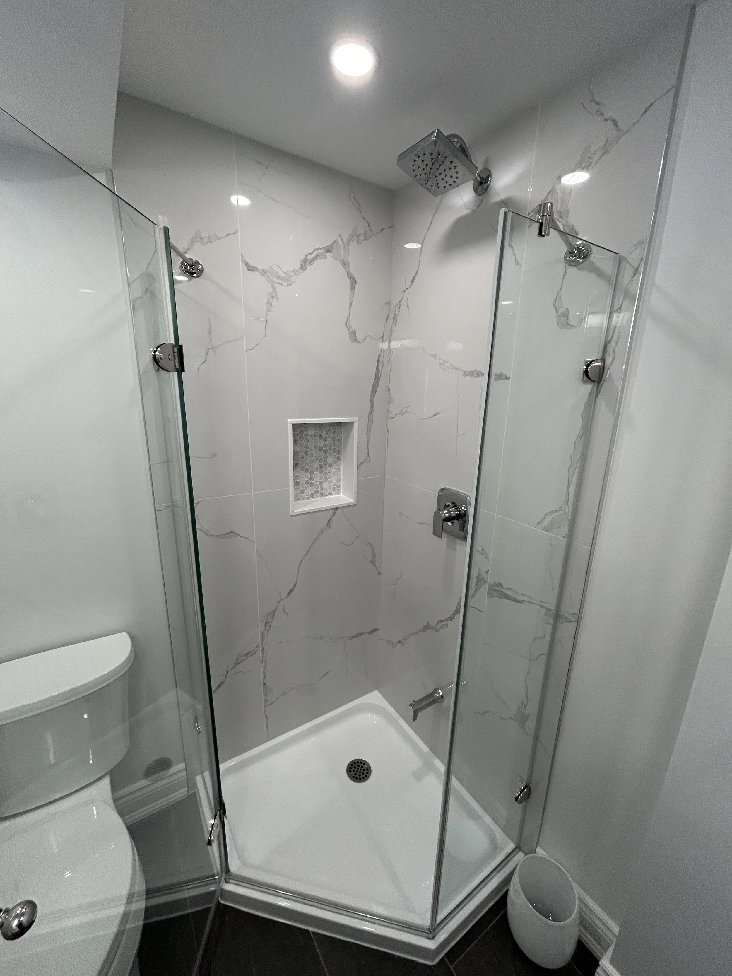 Shower stall with glass doors, marble wall tiles, a shower head, and a drain on the white shower floor.