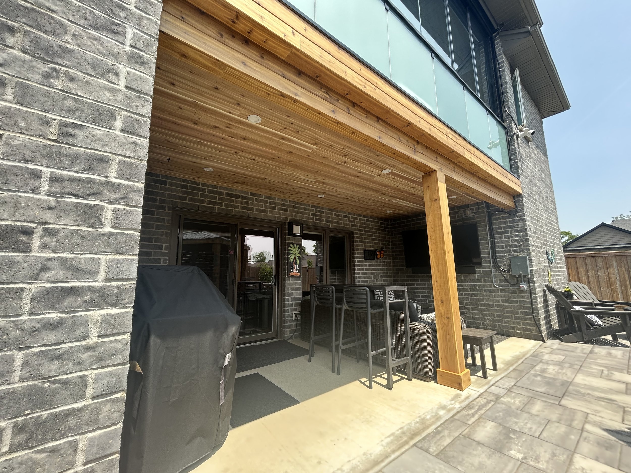 View of a modern house's backyard patio area with brick walls, wooden ceiling under a balcony, patio furniture, a barbecue grill, and a paved ground. A small flower pot is on a side table, and part of a wooden fence is visible in the background.