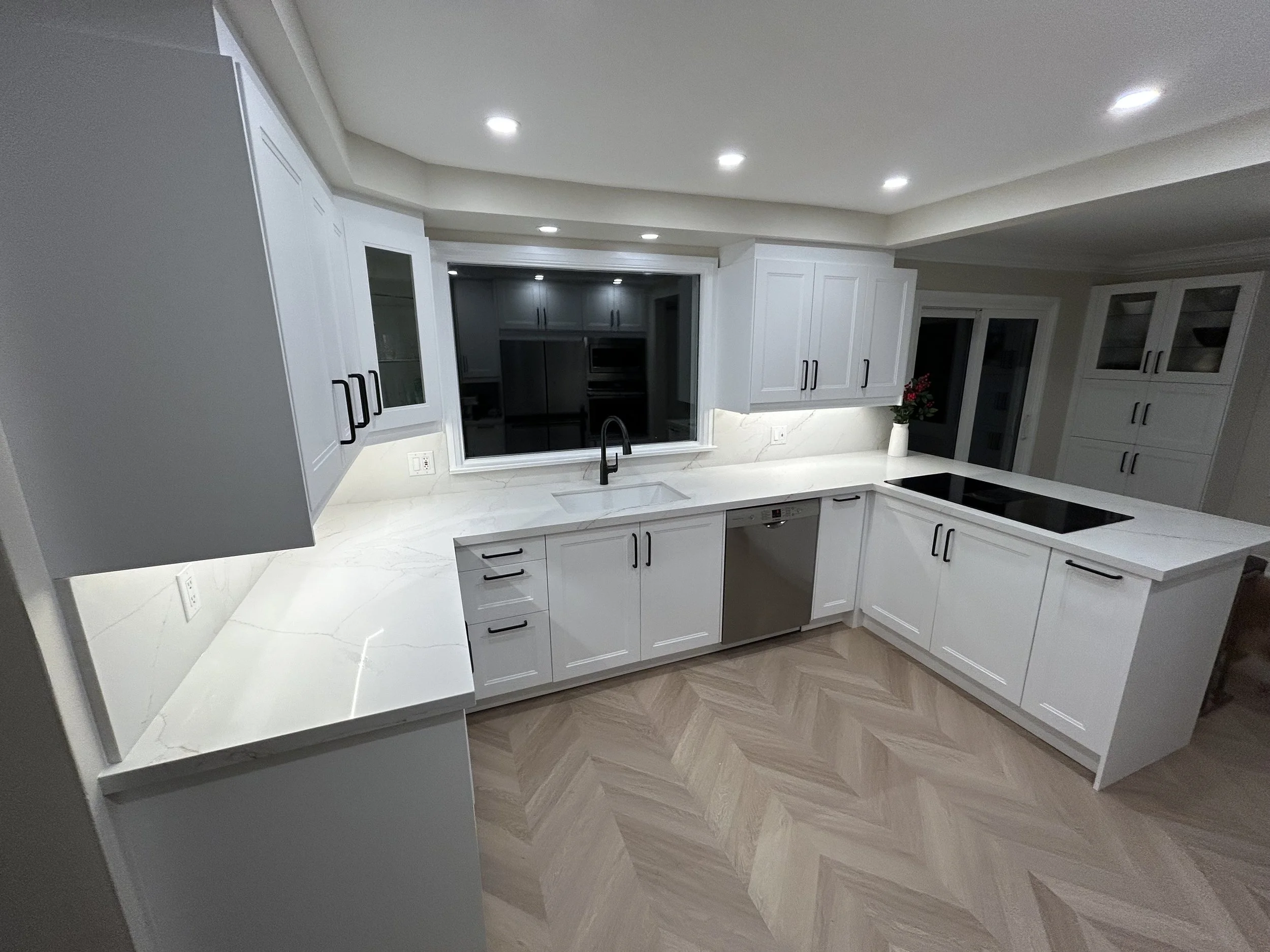 Modern kitchen with white cabinetry, marble countertops, and a window overlooking a dark room or outdoor space. Recessed ceiling lights and a potted flower are also visible.