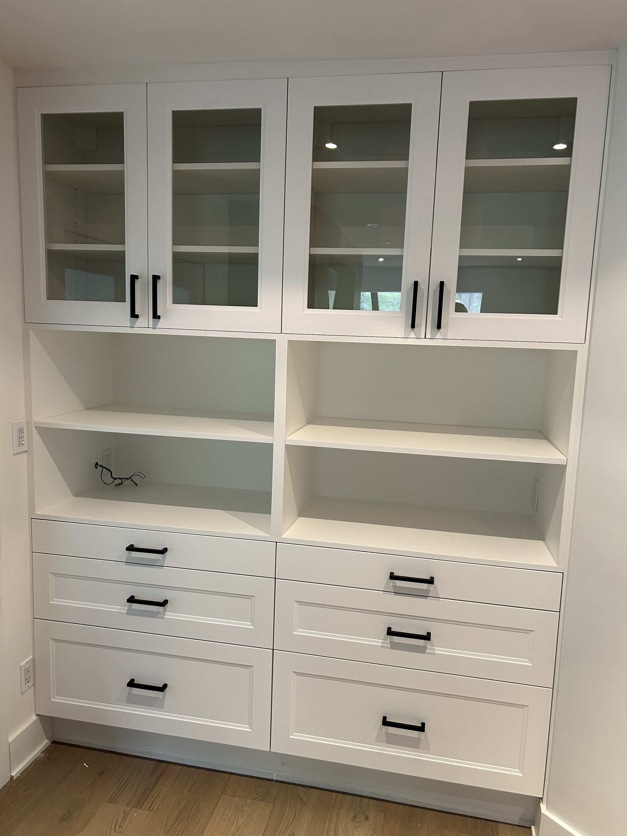 White built-in cabinet with glass-front upper section and multiple drawers below, black handles, in a room with wood floor.