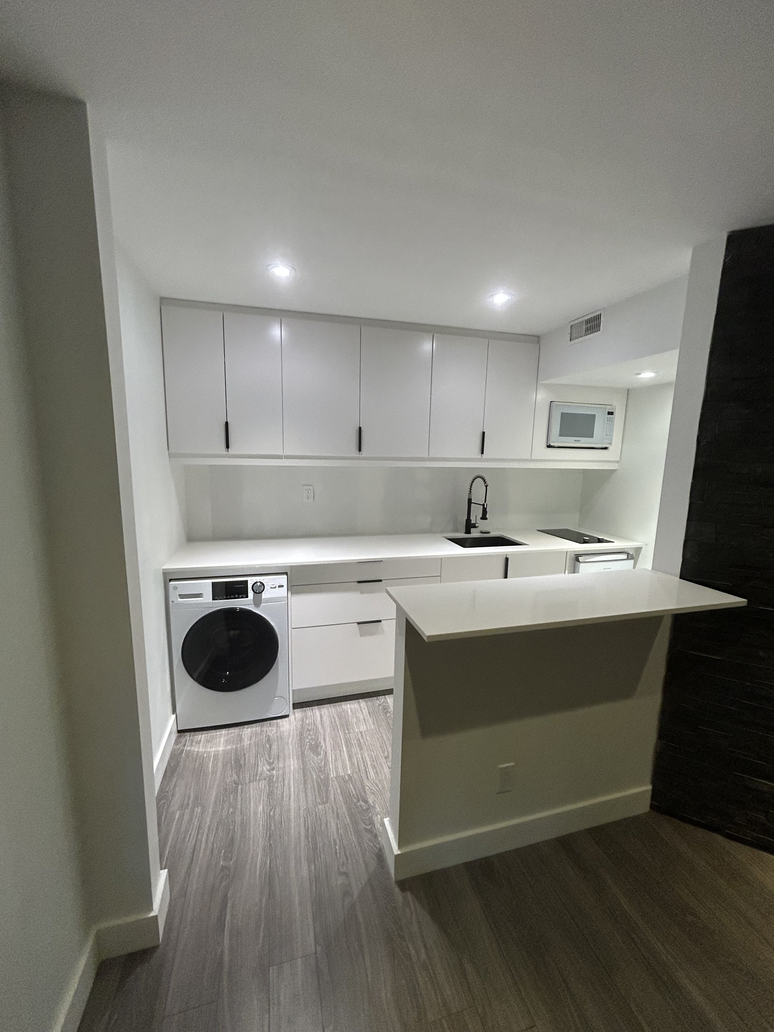Modern kitchen with white cabinets, black sink, microwave, and washer, featuring wood-look flooring and a breakfast bar.