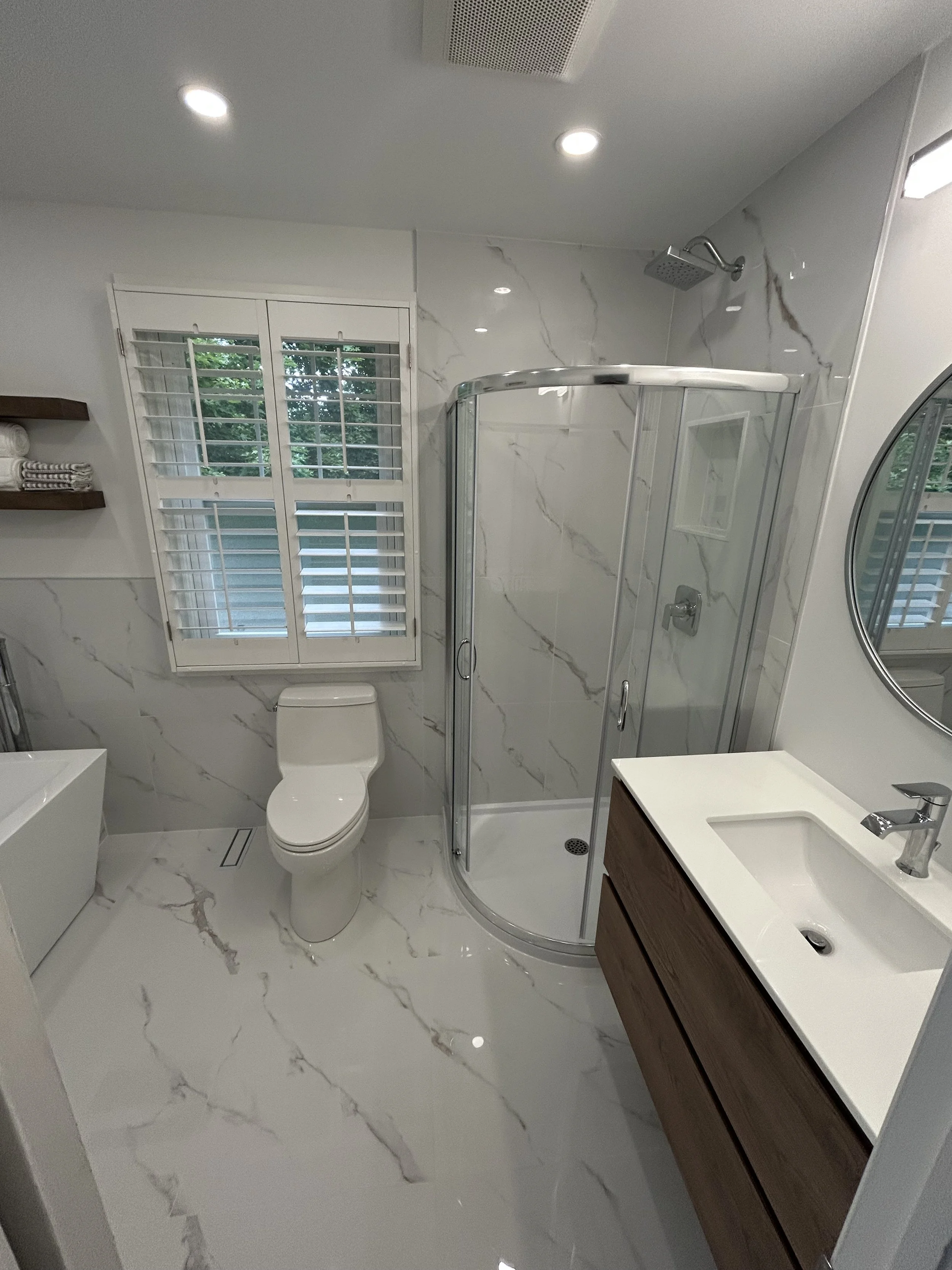 Modern bathroom with white marble floors and walls, a white toilet, a curved glass shower enclosure with a rain showerhead, a window with white plantation shutters, a wooden vanity with a white sink, and a round mirror.