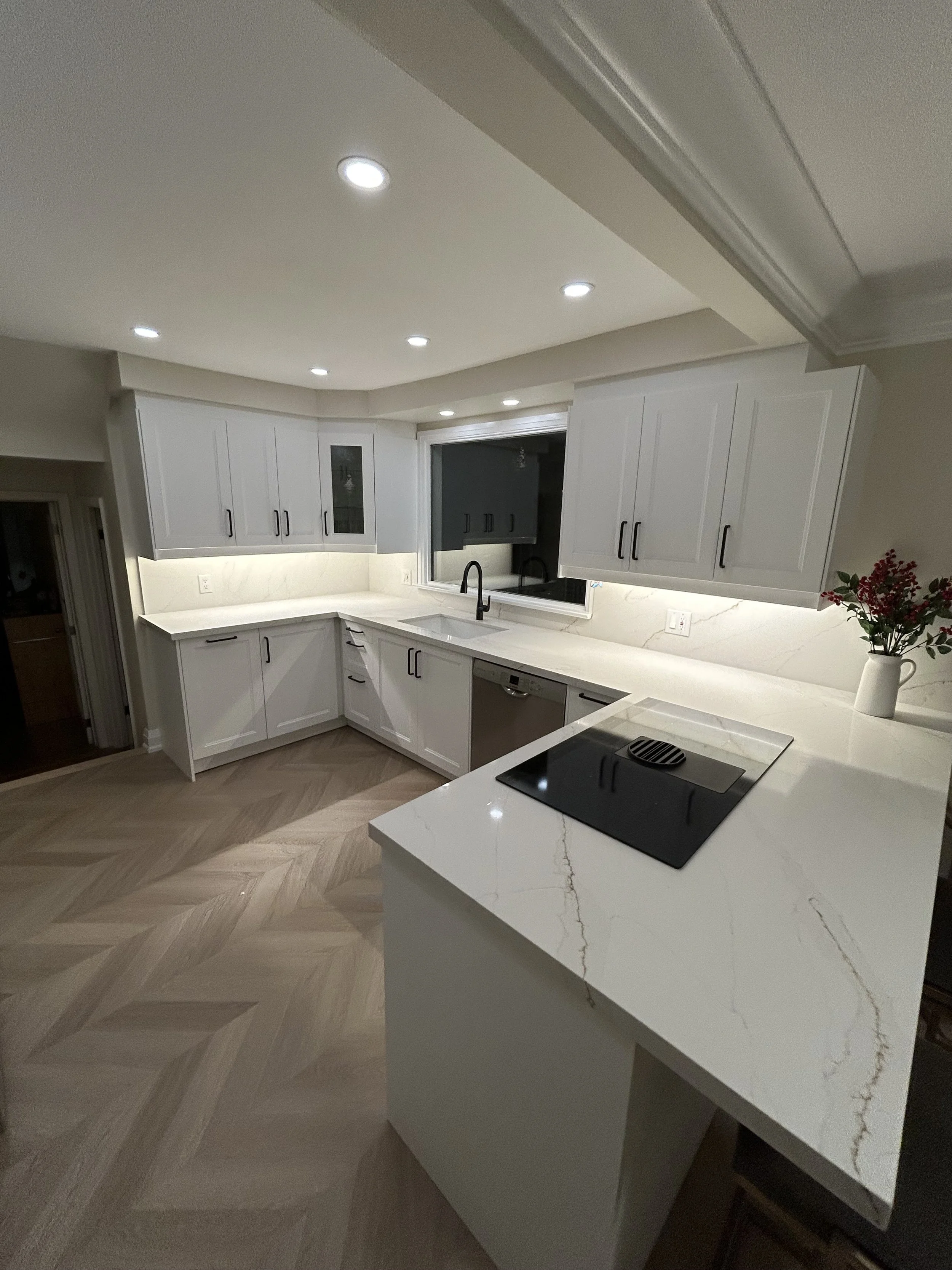 Modern kitchen with white cabinets, marble countertop, black faucet, built-in stovetop, and a vase with flowers on the counter, under ceiling spotlights.