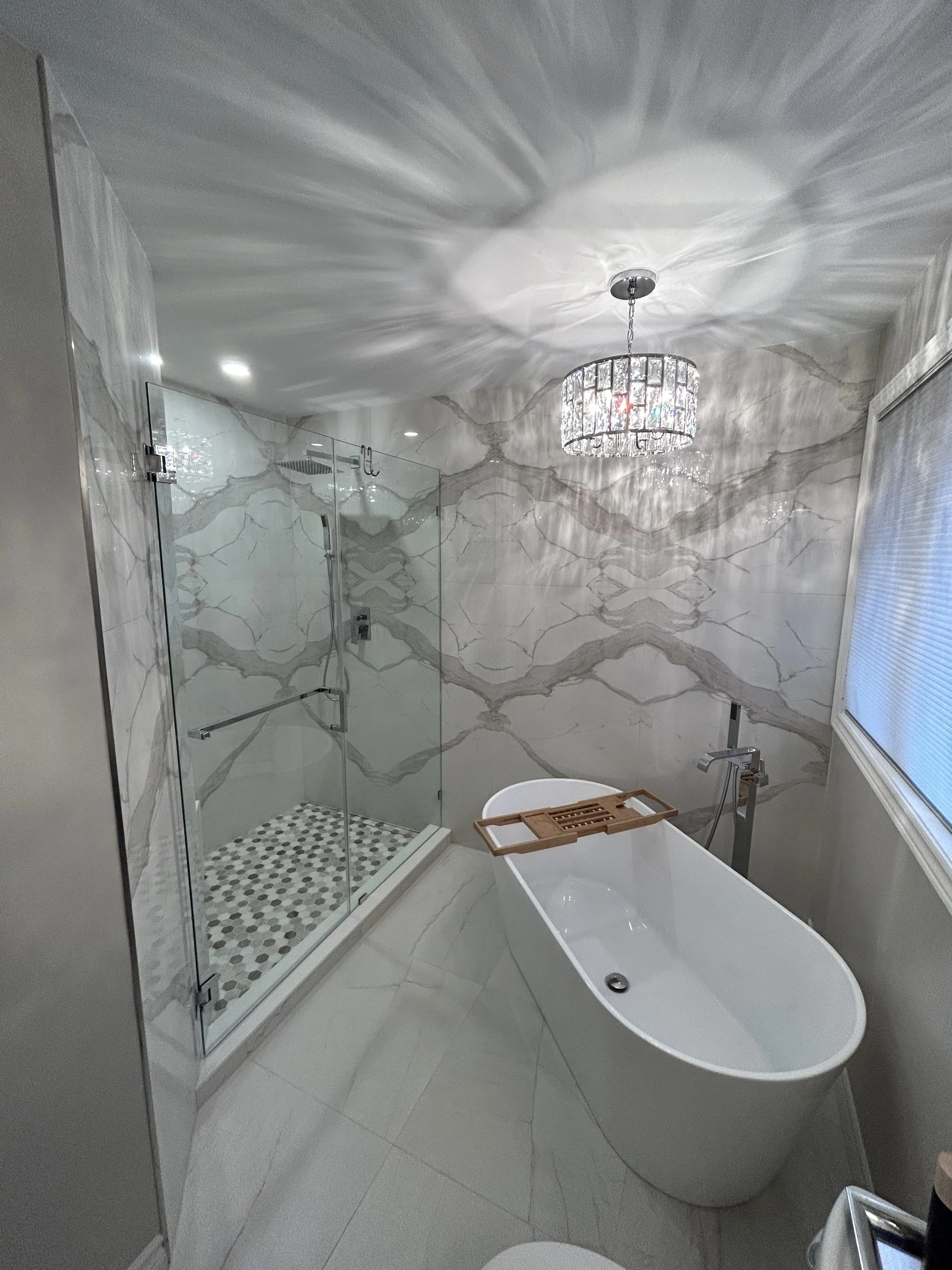 Modern bathroom with a glass shower enclosure, a freestanding bathtub with a wooden tray, marble wall and floor tiles, a chandelier casting light shadows on the ceiling, a window with closed blinds, and a small platform with a device on it.