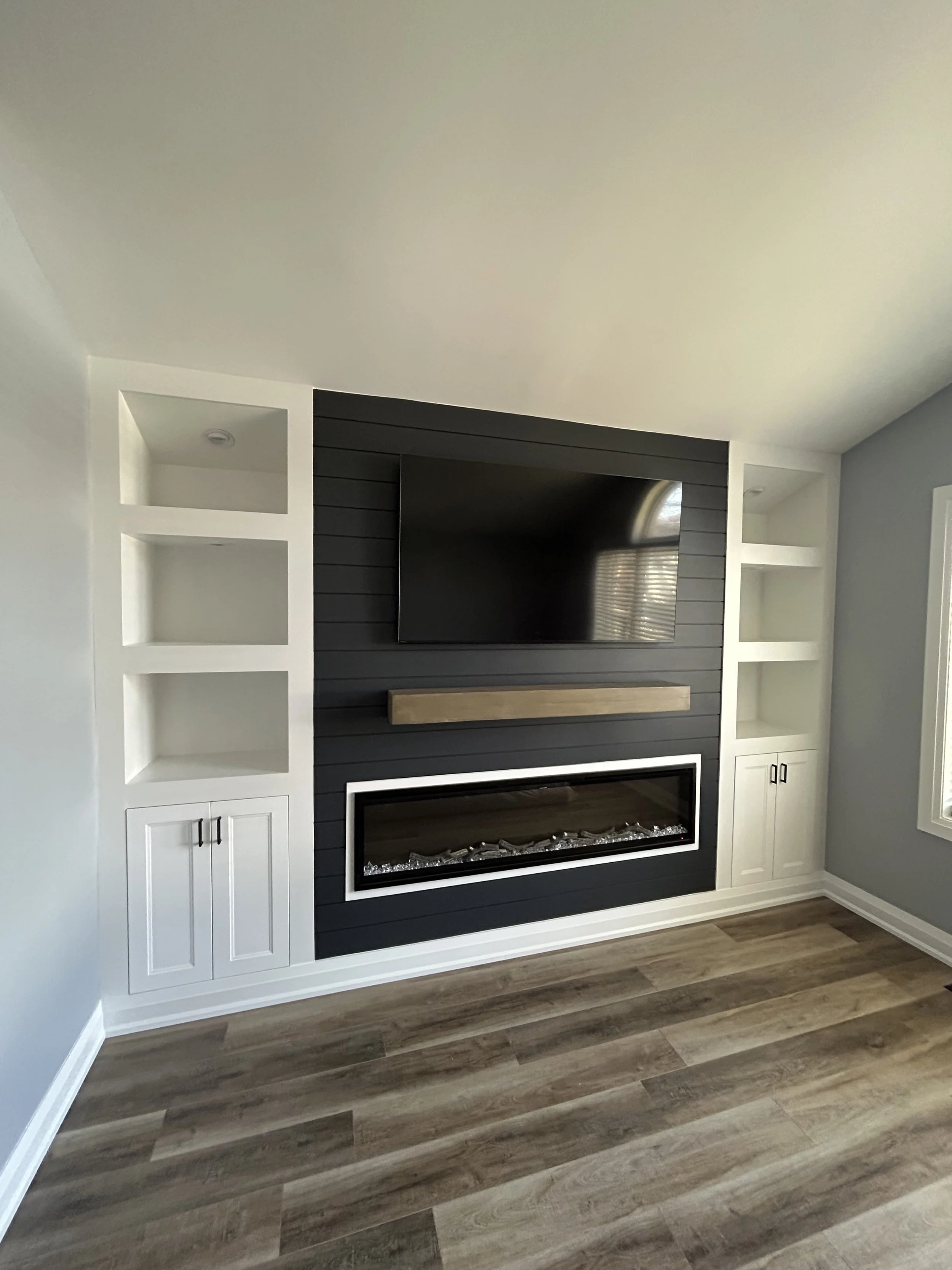 Living room fireplace with black shiplap surround, wood mantle, and built-in white cabinets with black handles on either side. Hardwood flooring and light-colored walls.