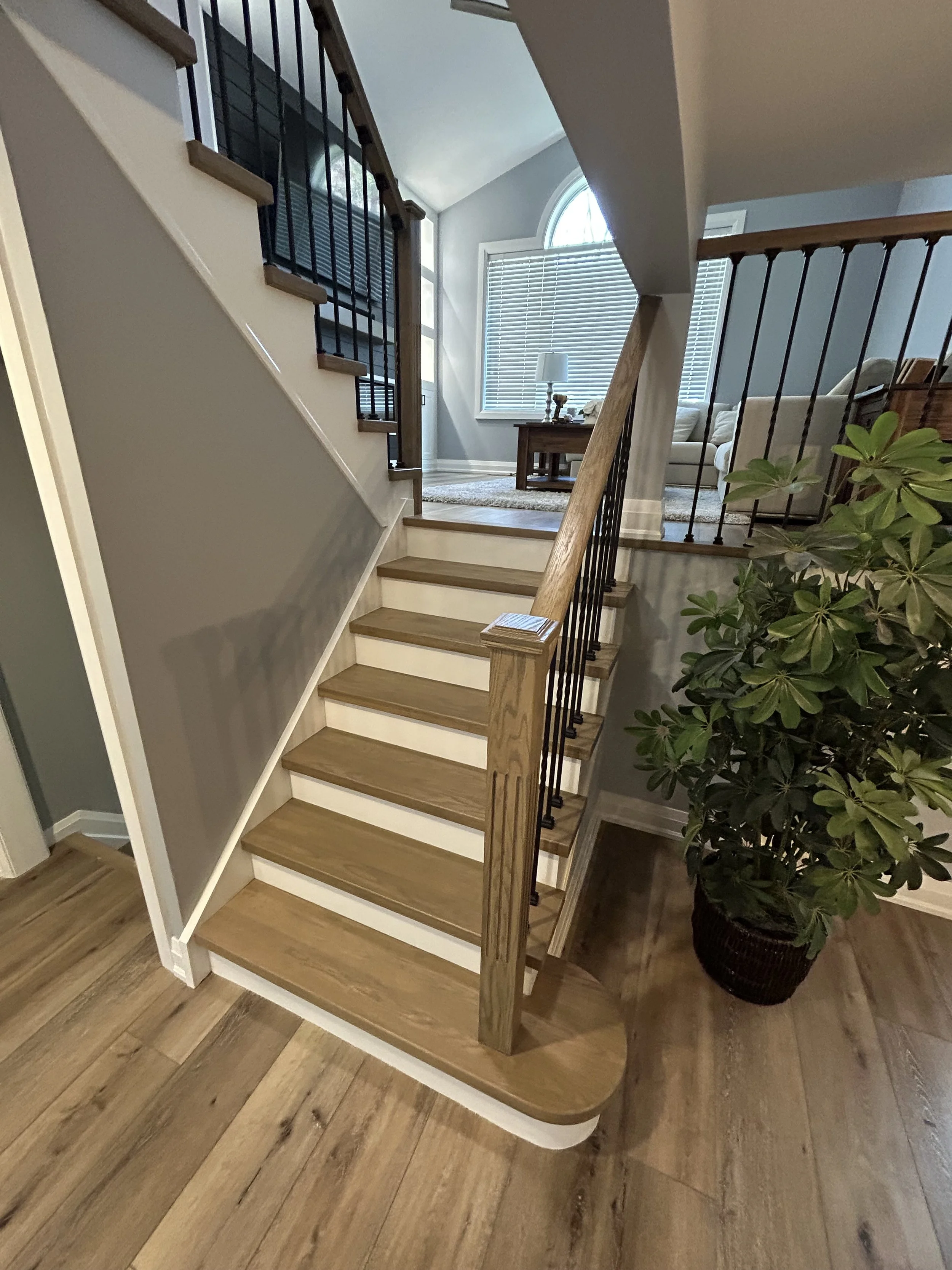 View of a staircase with wooden steps, metal railing, leading to a living room with a beige couch, a wooden table, a white lamp, and a large window with blinds. There is a large potted plant with green leaves next to the staircase. The floor is made of wooden planks.