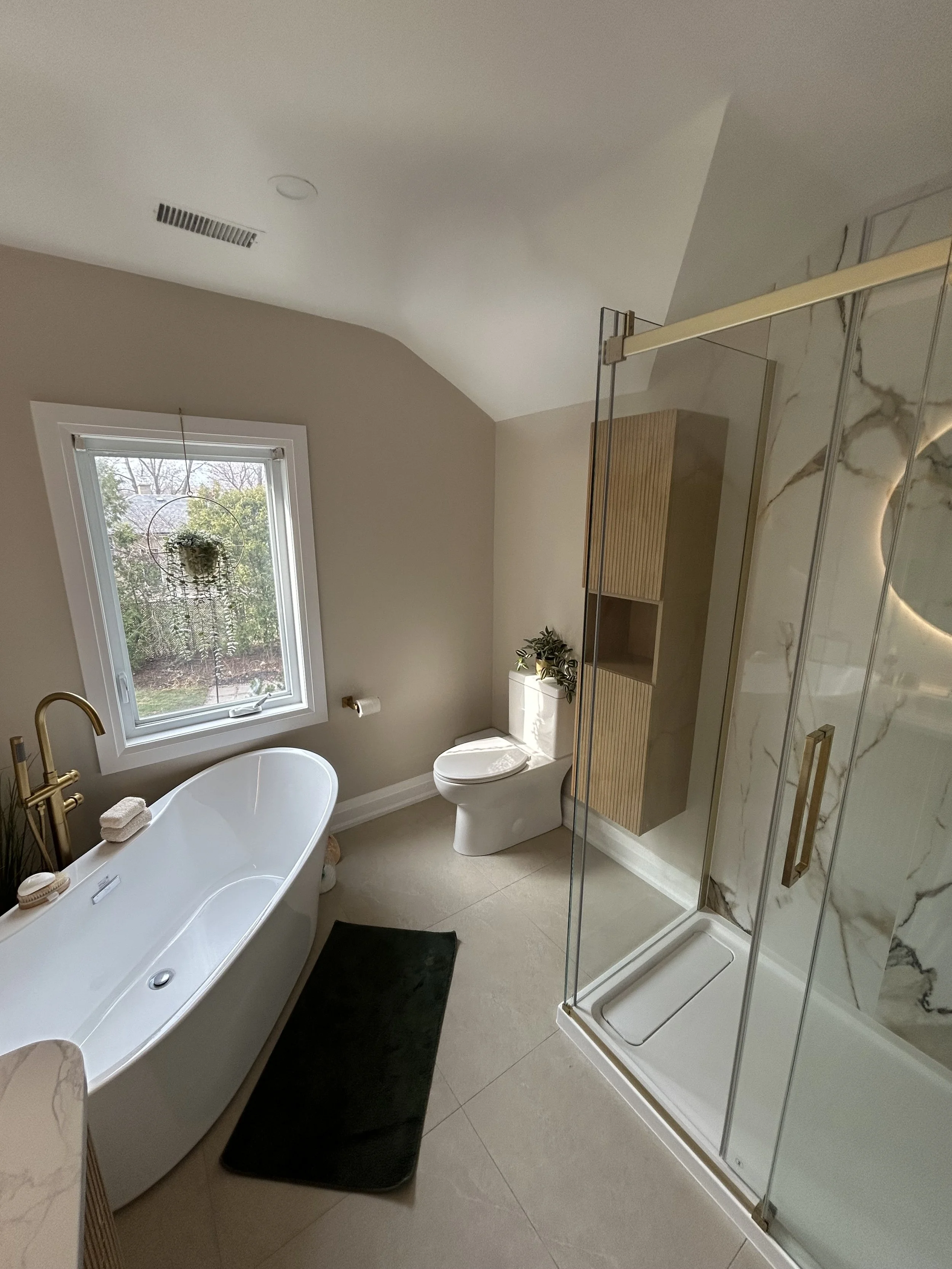 A modern bathroom with a freestanding bathtub, a toilet, a glass-enclosed shower, a window with a plant, and beige and marble decor.