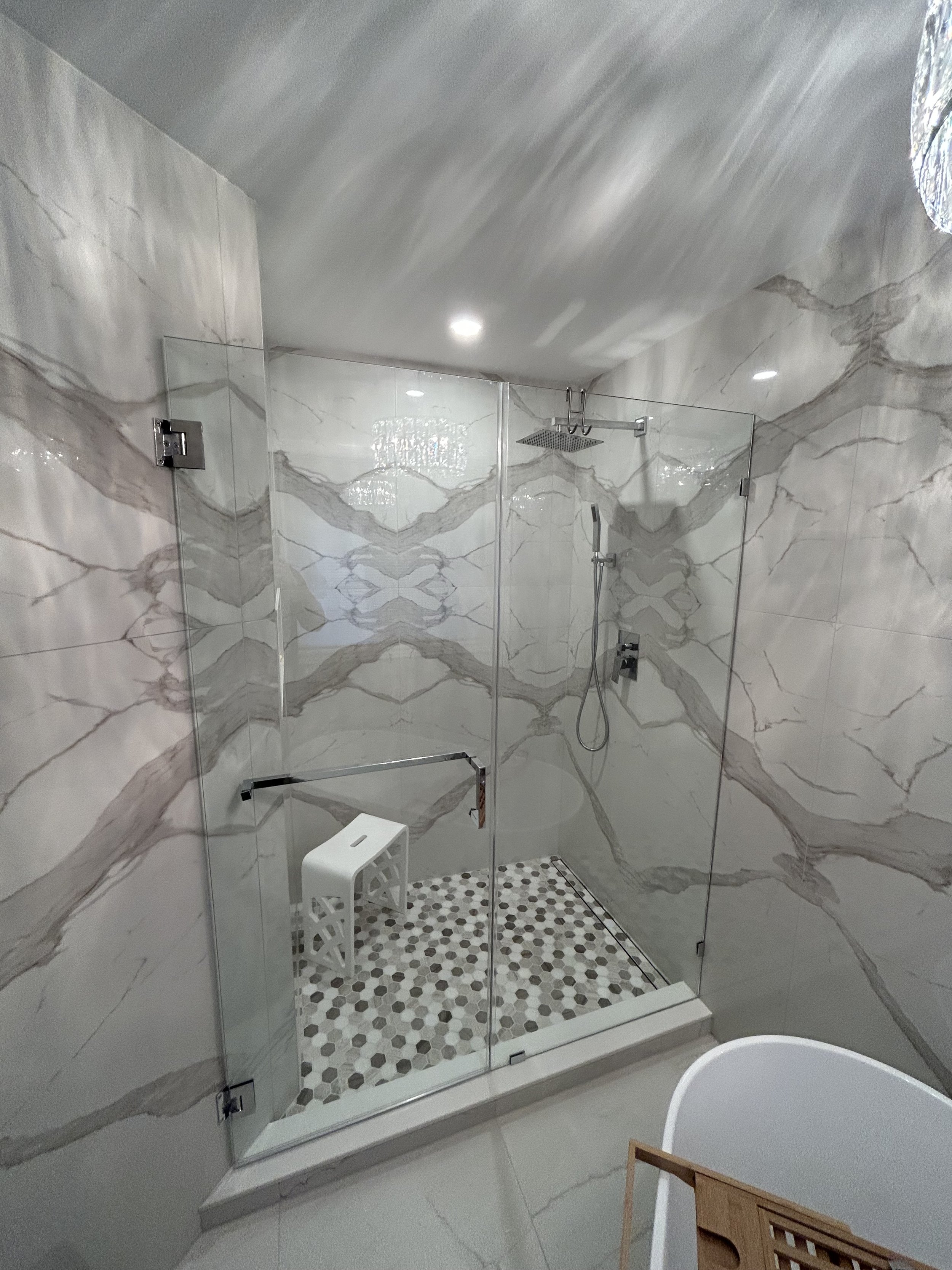 Modern walk-in shower with marble walls featuring gray and white veining, glass enclosure, rainfall showerhead, handheld shower, and small bench with patterned floor tiles in black, white, and gray.